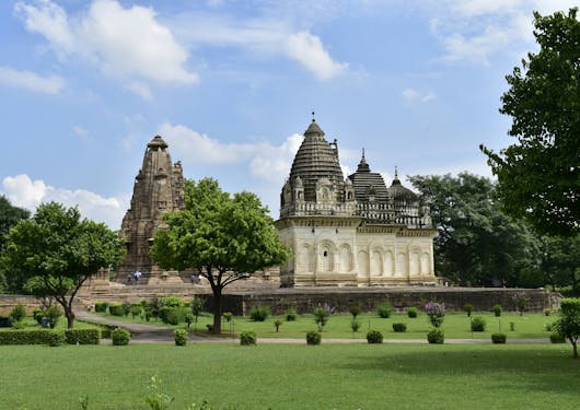The Khajuraho Temple in India is shown from a distance, with a perfectly manicured, bright green garden in front of it and some luscious green trees to the right, behind it, and a couple to the left. The sky is partially cloudy.