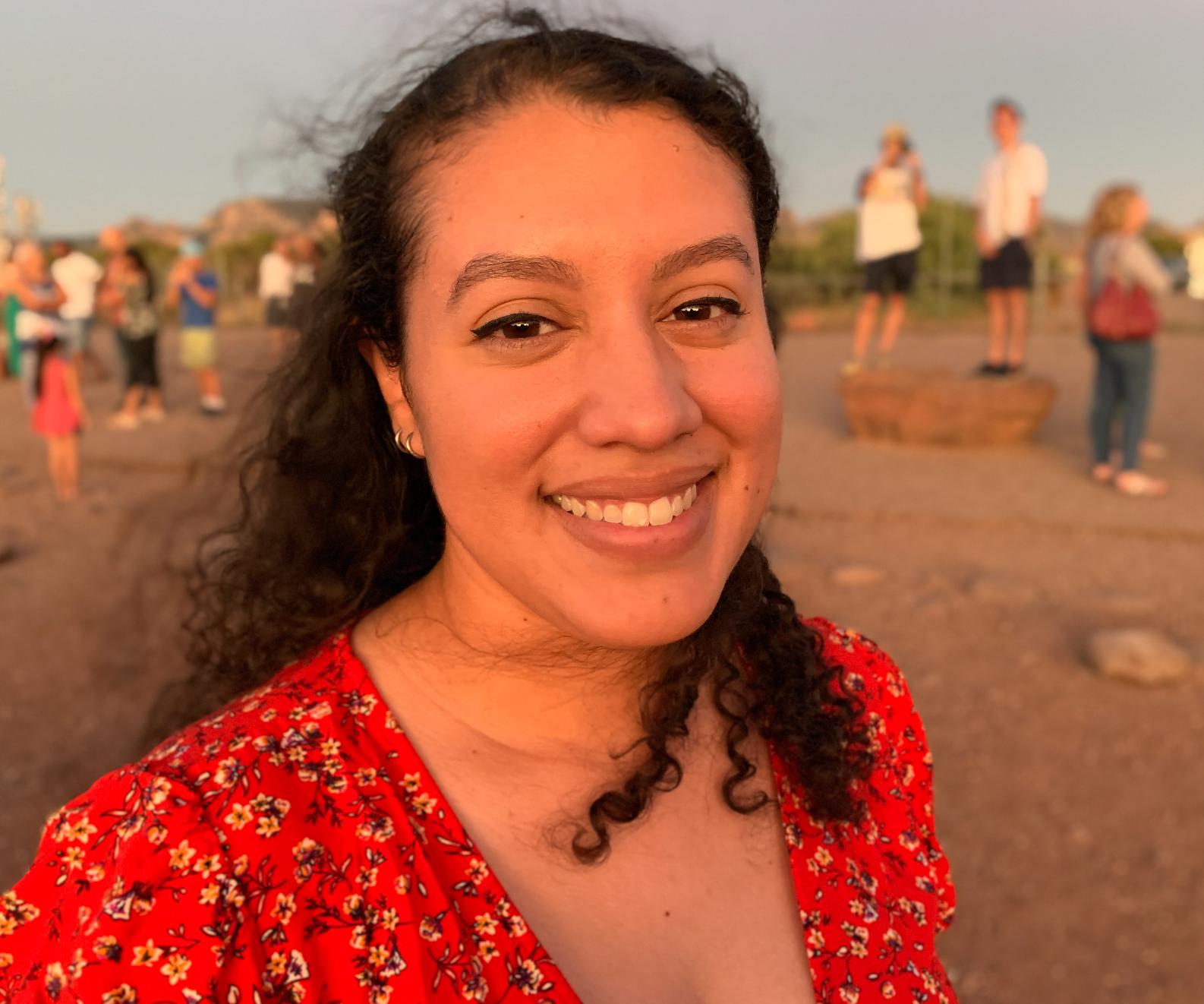 A headshot of a young woman with long, dark, curly and coarse hair, pulled half back behind her, smiling at the camera. She's standing on what appears to be a beach with other people in the background.