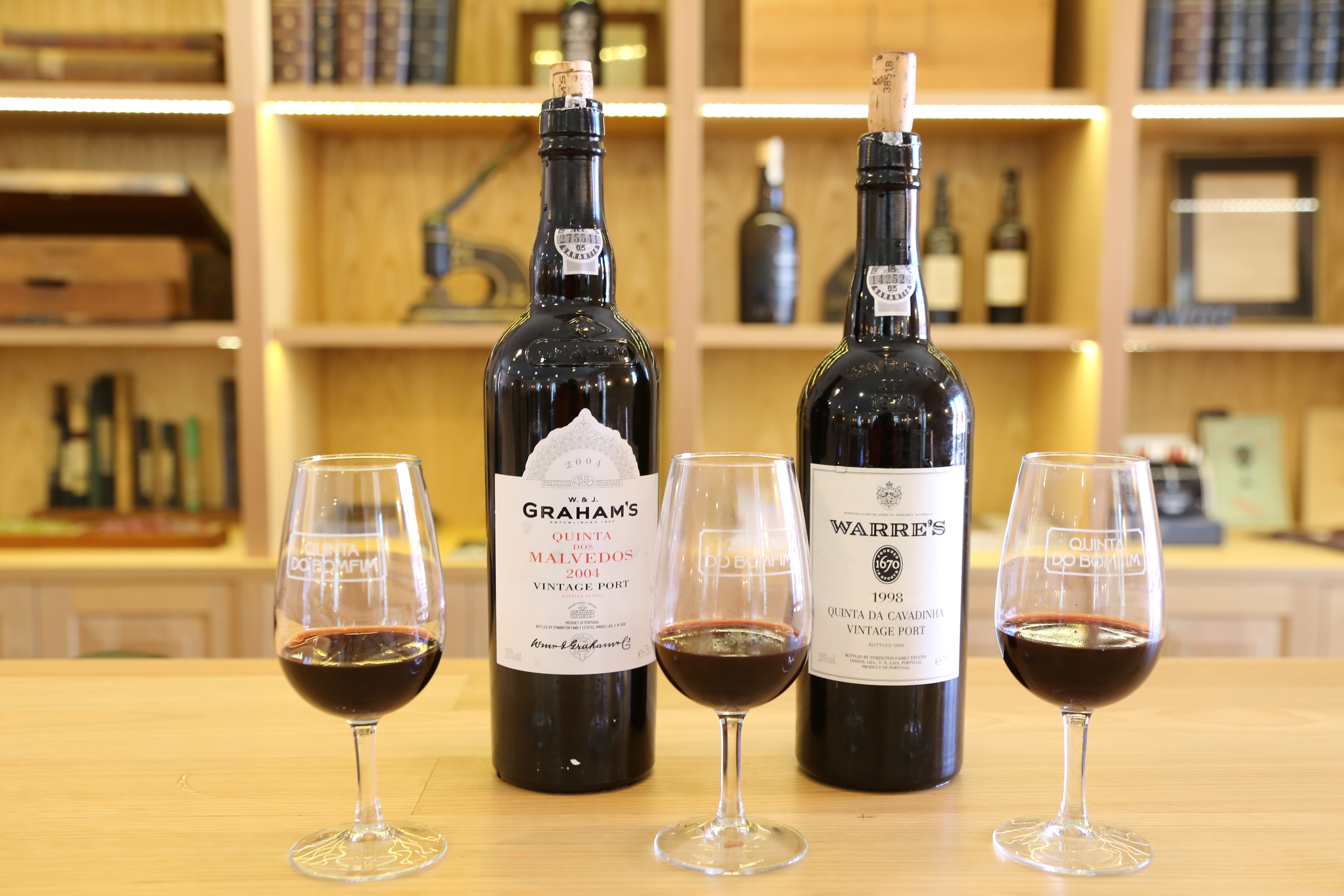 2 wine bottled of Port wine sit behind 3 wine glasses filled with red wine on a beige countertop. Shelves with wine bottles and books are in the background.