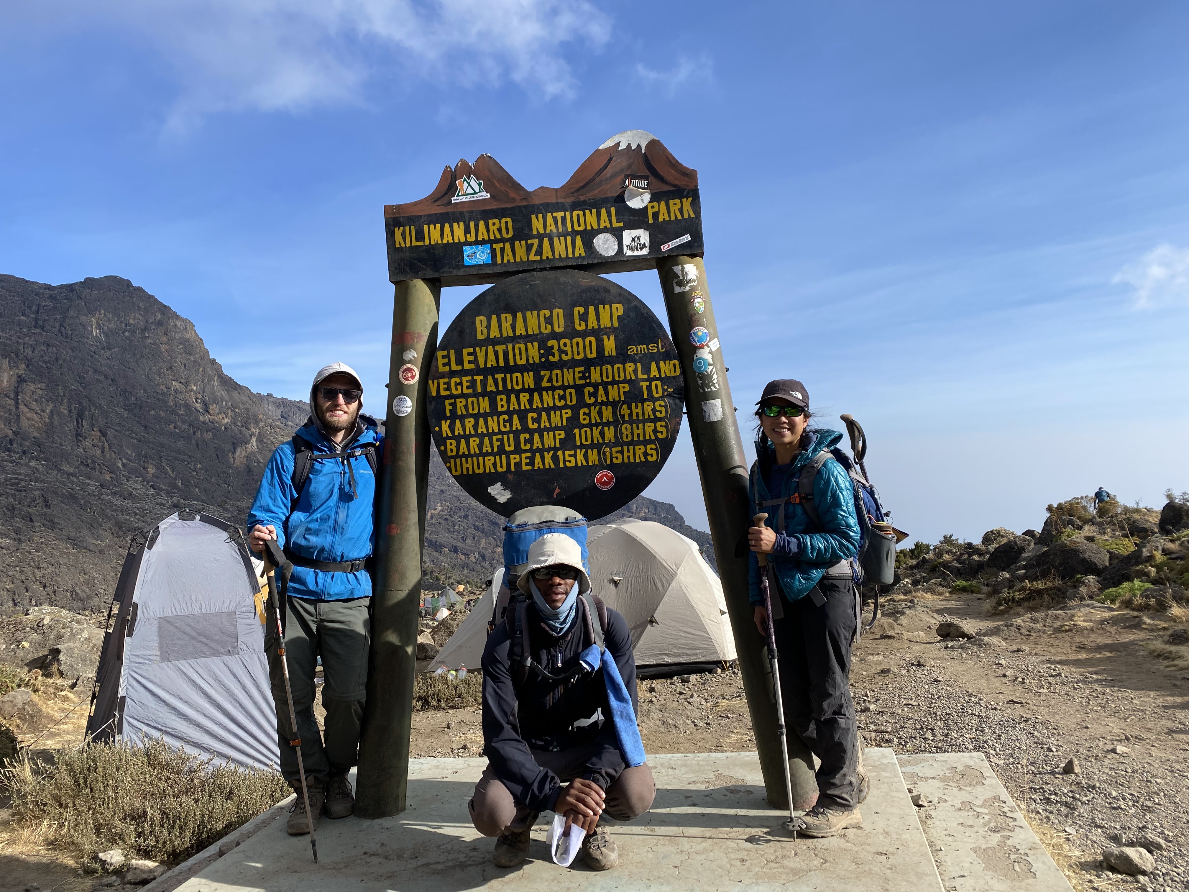 Two climbers, a man and a woman, prepare to trek Mount Kilimanjaro in Tanzania with their climb starting at the Baranco Camp. The camp signpost is on wooden and metal materials with yellow lettering. there are some tents in the background, the couple stands to either side of the sign, and the guide squats in front of the sign.