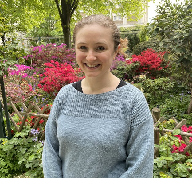 A Caucasian woman with blond hair and a blue knit sweater face the camera smiling, standing in front of a lush green background with clusters of red and majenta flowers.