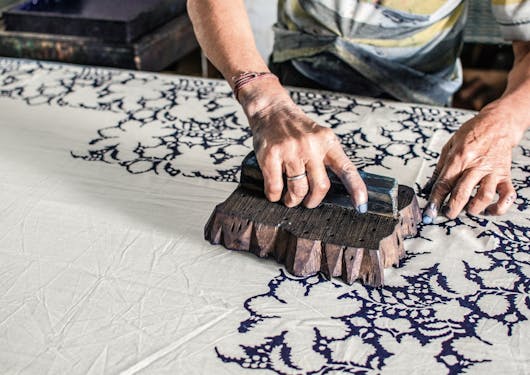 A block printer in India demonstrates his traditional, artisanal craft on the Golden Triangle Cultural Tour. The block is wooden and it is pressing a dark dye into a beige textible. The artisan's naiels are blue and only their forearms and hands are visible.