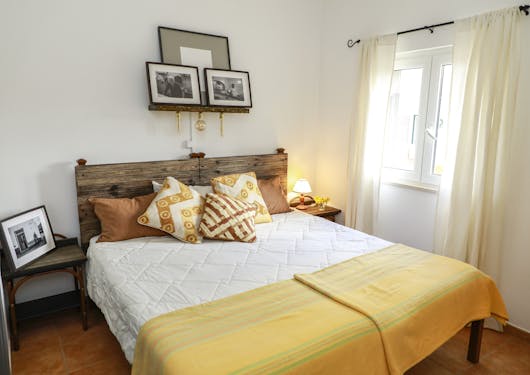 A bedroom at the sustainable Portugal yoga retreat center shows a large bed with yellow and brown pillows and a matching yellow throw blanket. Picture frames are up above and a window with light coming through is on the right hand side. There's another picture frame to the left of the bed and a small lamp lit on the right hand side.