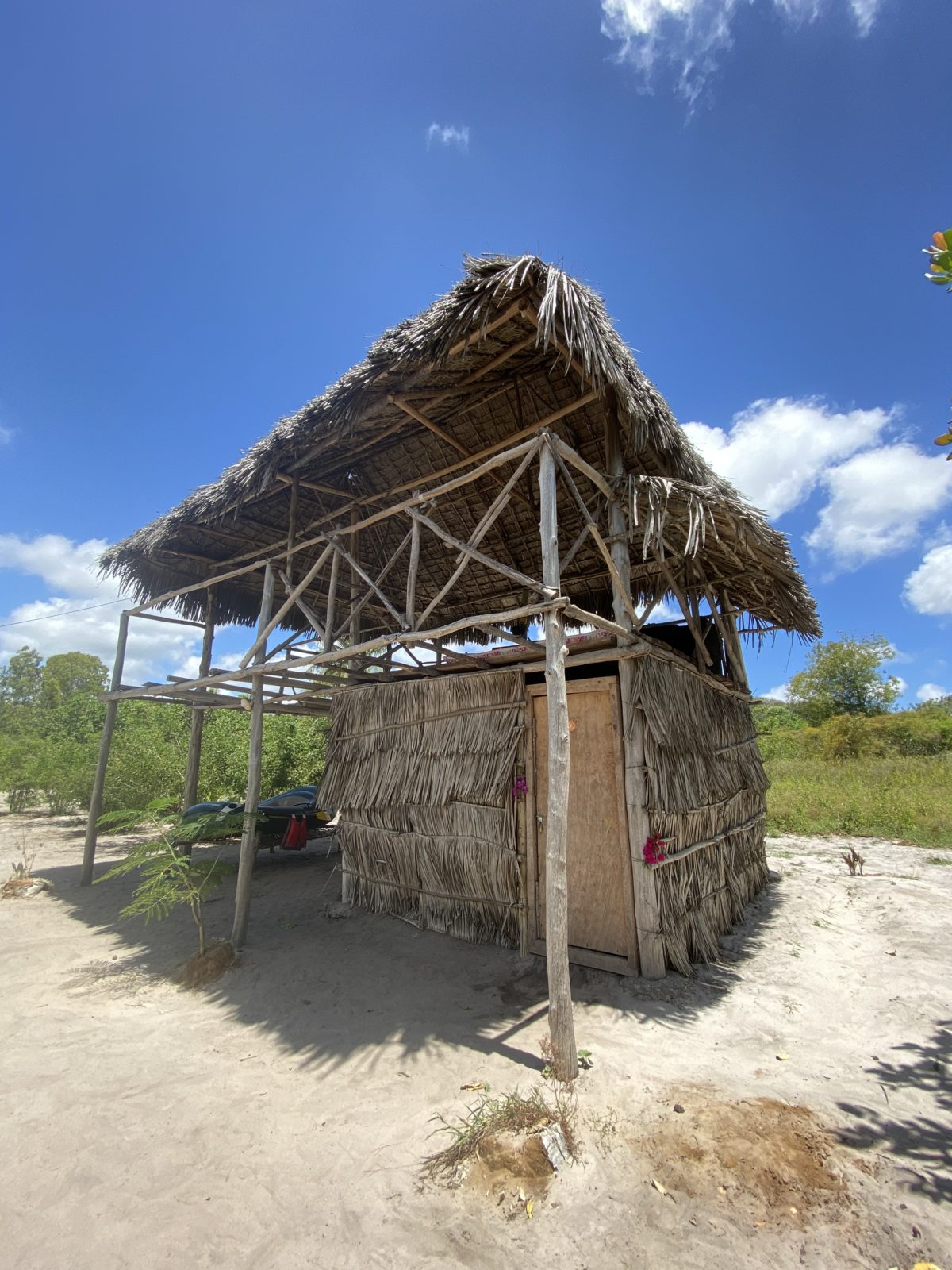 A 'makuti' traditional thatched roof stilted structure is shown in Kenya in the Mida Creek Community Village. There is greenery in the background and the sky is blue with a couple of white clouds.