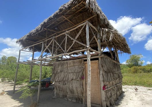 A 'makuti' traditional thatched roof stilted structure is shown in Kenya in the Mida Creek Community Village. There is greenery in the background and the sky is blue with a couple of white clouds.