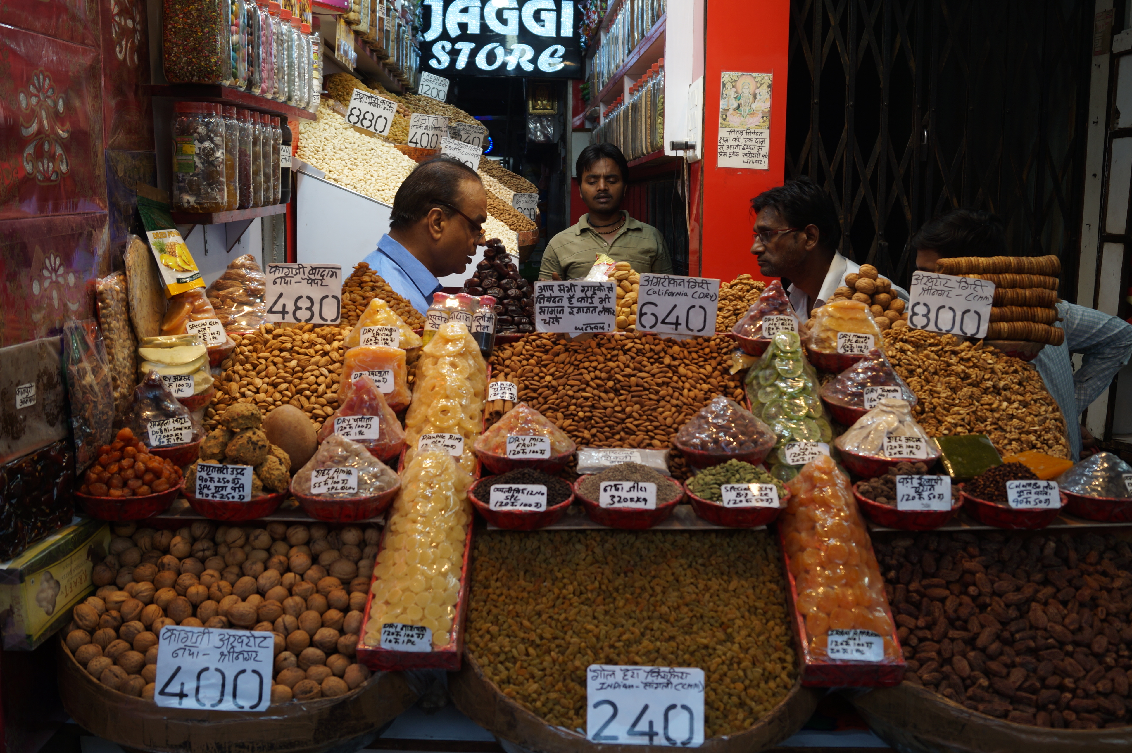 Three Indian men stand behind a market stall booth in Old Agra, India. Colorful nuts and dried fruit are laid out in neat piles and rows. This is something you can see on the Golden Triangle Cultural Tour of India.