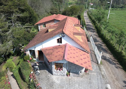 The Villa Doris Country Lodge, a sustainable, eco-lodge near the village of Nona in Ecuador sits in the center of the frame, with tall, green trees to the left and a grassy field to the right.