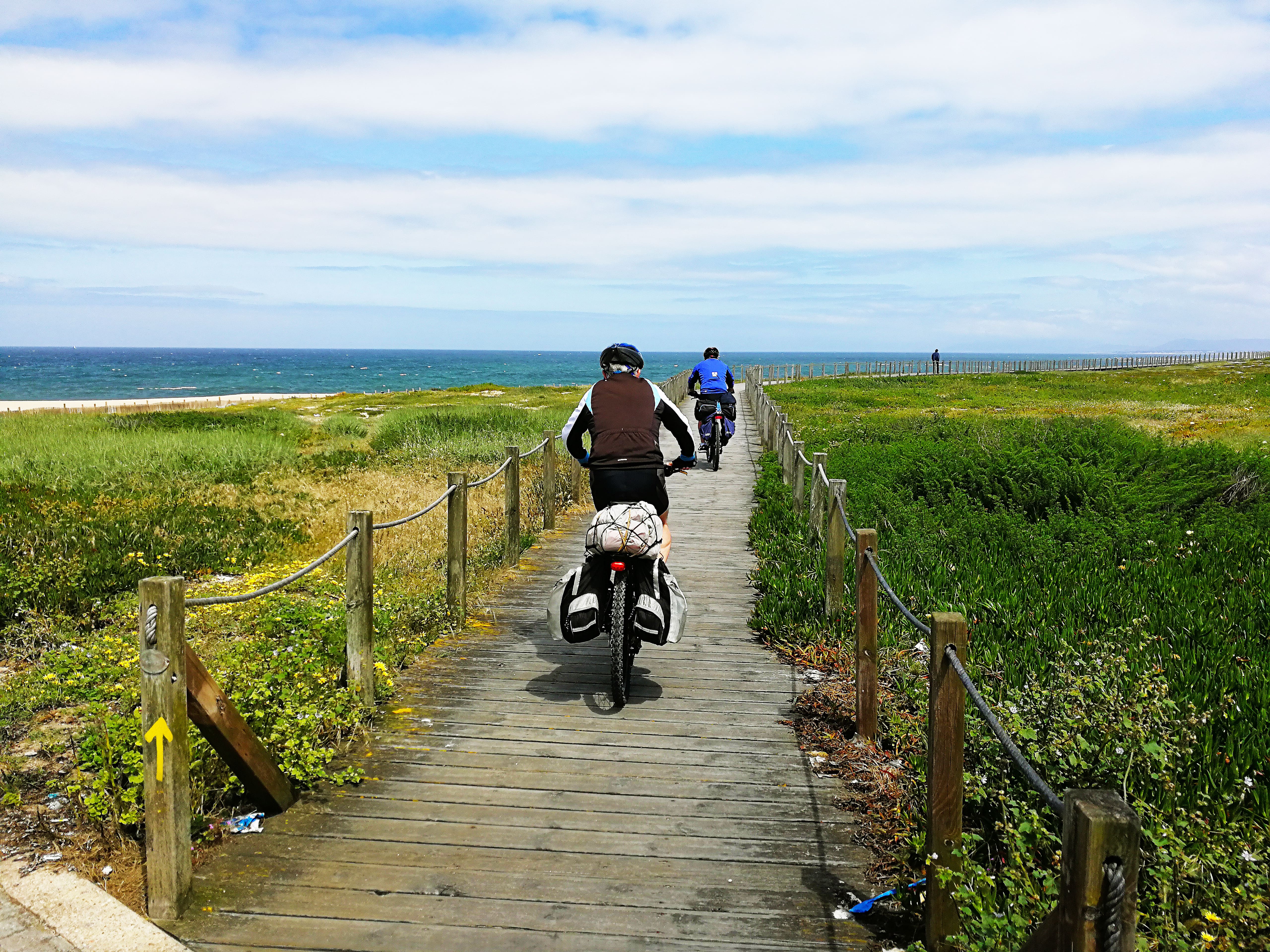 2 bikers cycle along a wooden boardwalk near the Atlantic Coast in northern Portugal under a sunny yet cloudy sky.