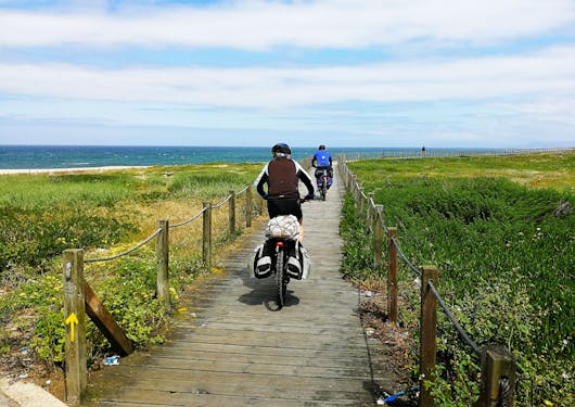 2 bikers cycle along a wooden boardwalk near the Atlantic Coast in northern Portugal under a sunny yet cloudy sky.
