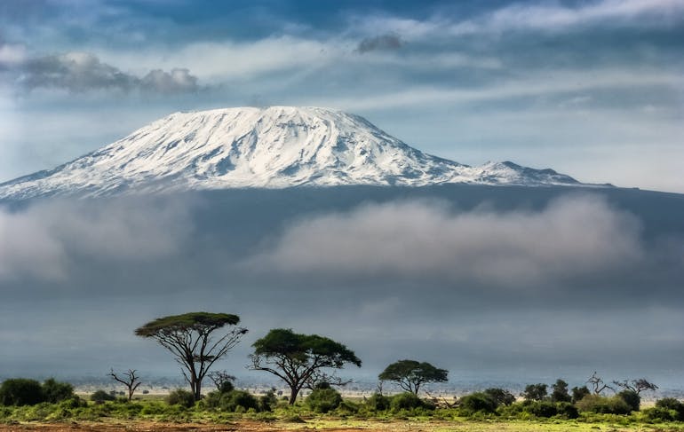 Mt Kilimanjaro in Tanzania shows a snow-capped peak above clouds; the desert is in the foreground with a few green trees.