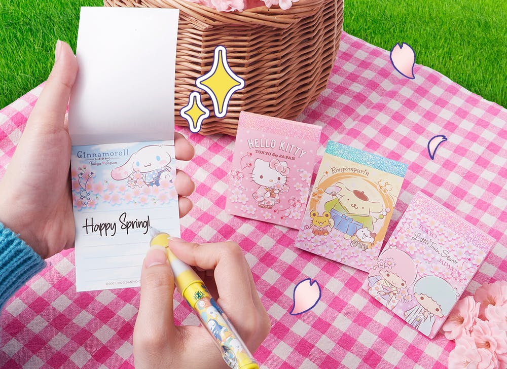 A picture of someone holding and writing in a Sanrio cherry blossom note pad featuring Hello Kitty, Pompompurin, and the Little Twin Stars Kiki and Lala. 