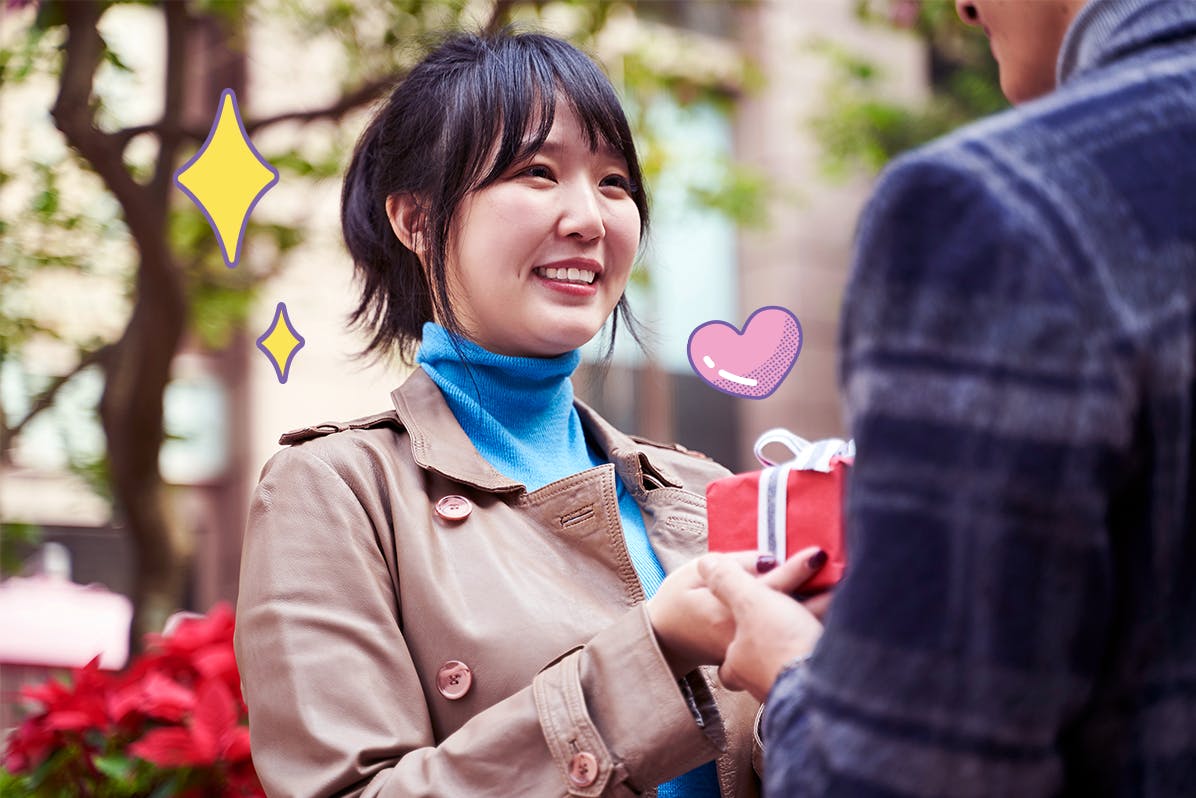 A picture of a young woman handing a red gift box to a man in front of her while smiling.
