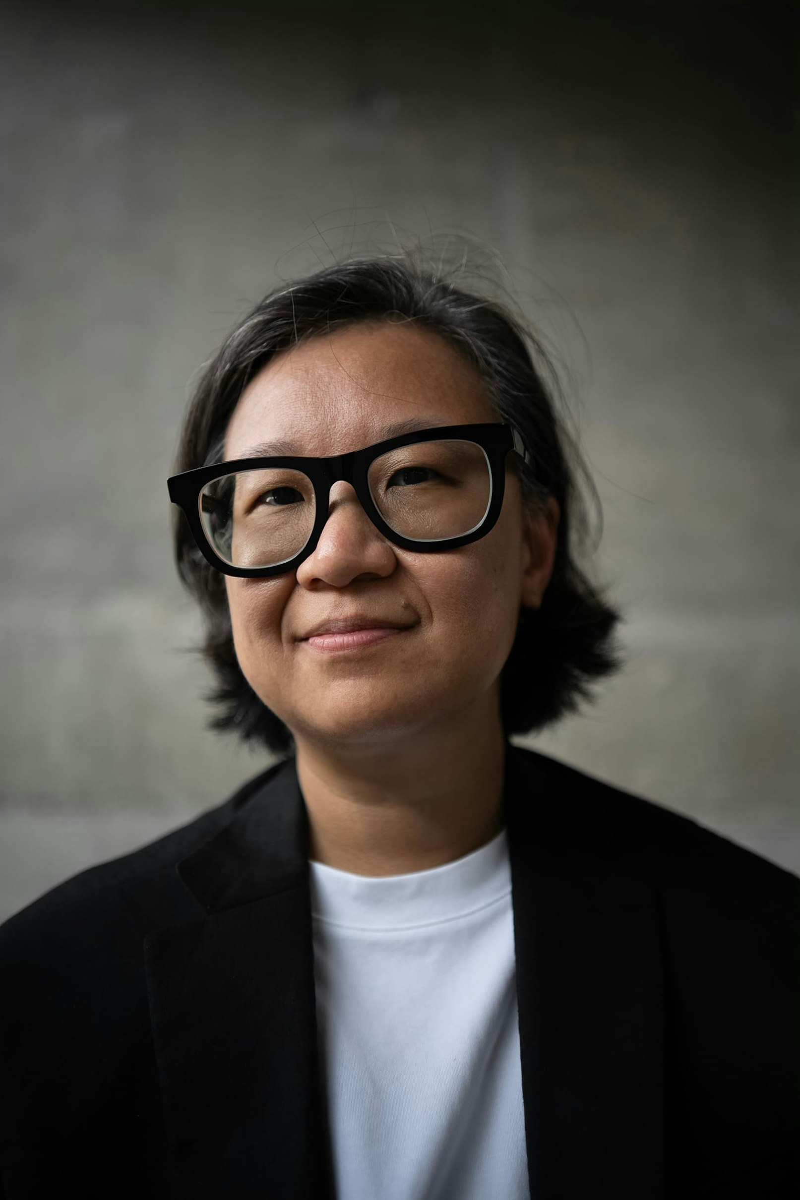 Connie is looking in the camera. She is wearing a white T-shirt and a black blazer. She has black, straight hair and wears bold black glasses. 