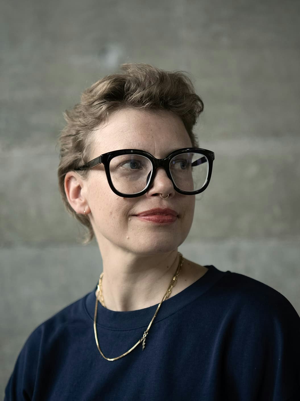 Denize is looking to the left, wearing a navy t-shirt and two gold necklaces, one with a lightening bolt. She has short light brown hair and bold black glasses.