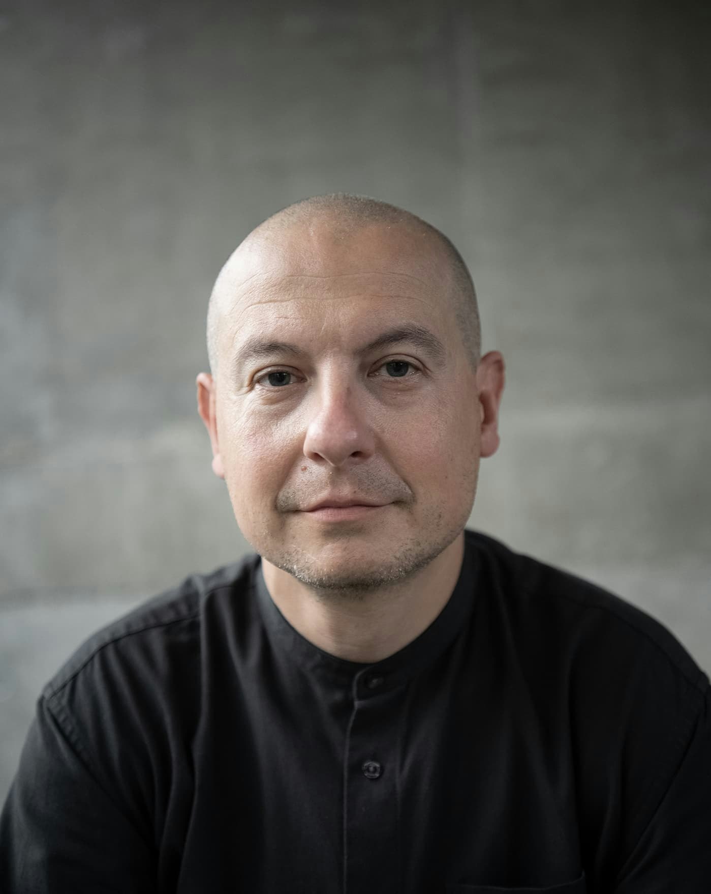 Diego is looking in camera wearing a dark green shirt. His head is shaved.