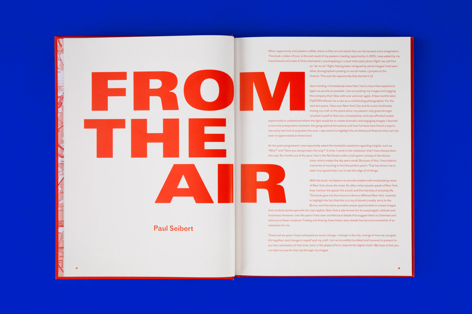 The book is open to a double spread with bright orange-red text on white ground. In bold large letters, it says FROM THE AIR. The book is photographed against a cobalt blue seamless.