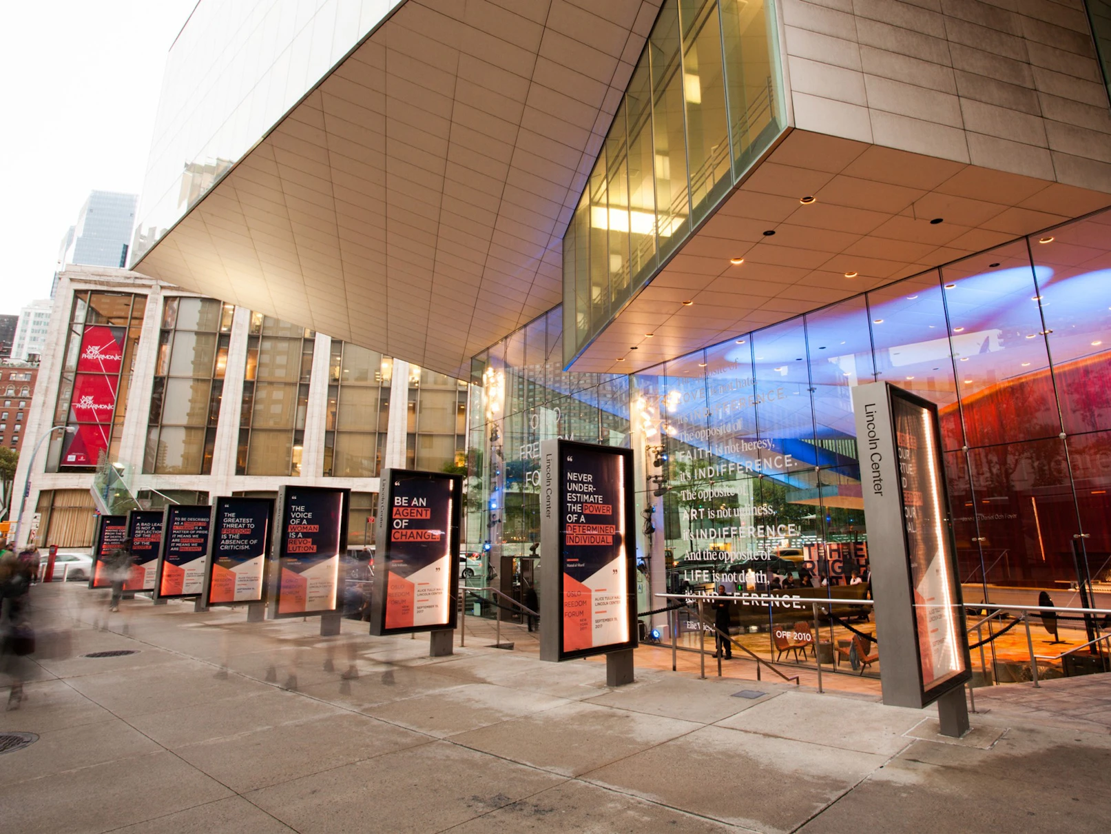 Image shows the exterior of a contemporary building with glass paneled walls. There is a row of outdoor signage that is covered in navy blue, coral, and white geometric graphics and text. There are people walking by and gathering near the signage. 