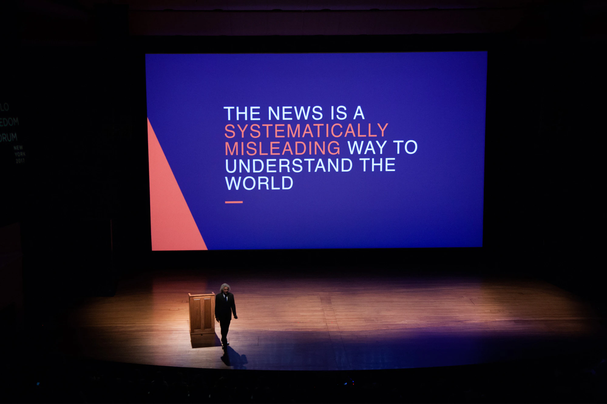 Image shows a person on a stage with wooden floors. There is a large screen behind them that shows the words THE NEWS IS A SYSTEMATICALLY MISLEADING WAY TO UNDERSTAND THE WORLD in white and orange letters on a blue background. There is a triangle of coral in the lower left corner of the screen.