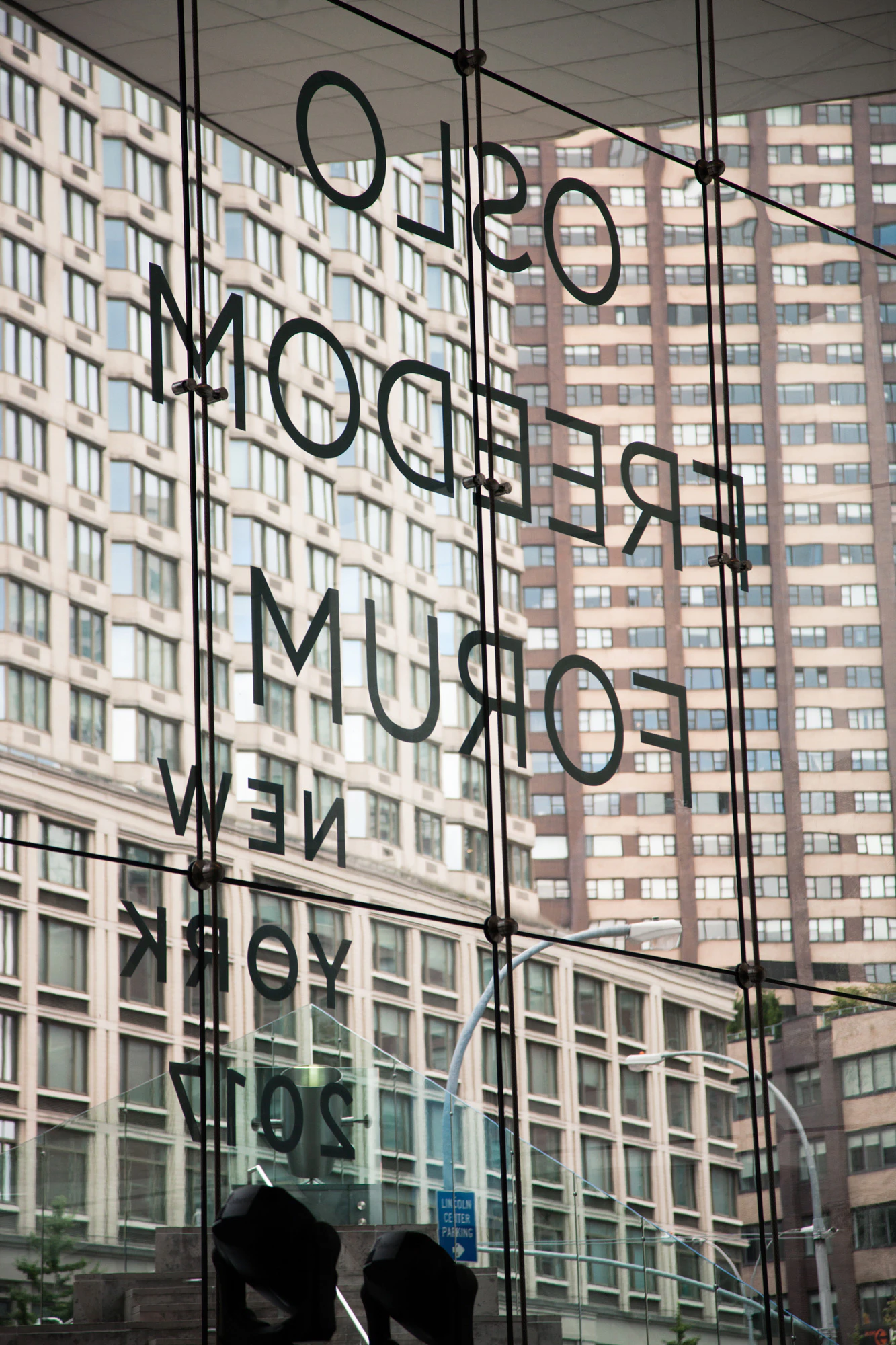 Image shows the words OSLO FREEDOM FORUM NEW YORK 2017 in decal letters on a window. The words are backwards because it is taken from inside the building. Through the window are multi-story city buildings.
