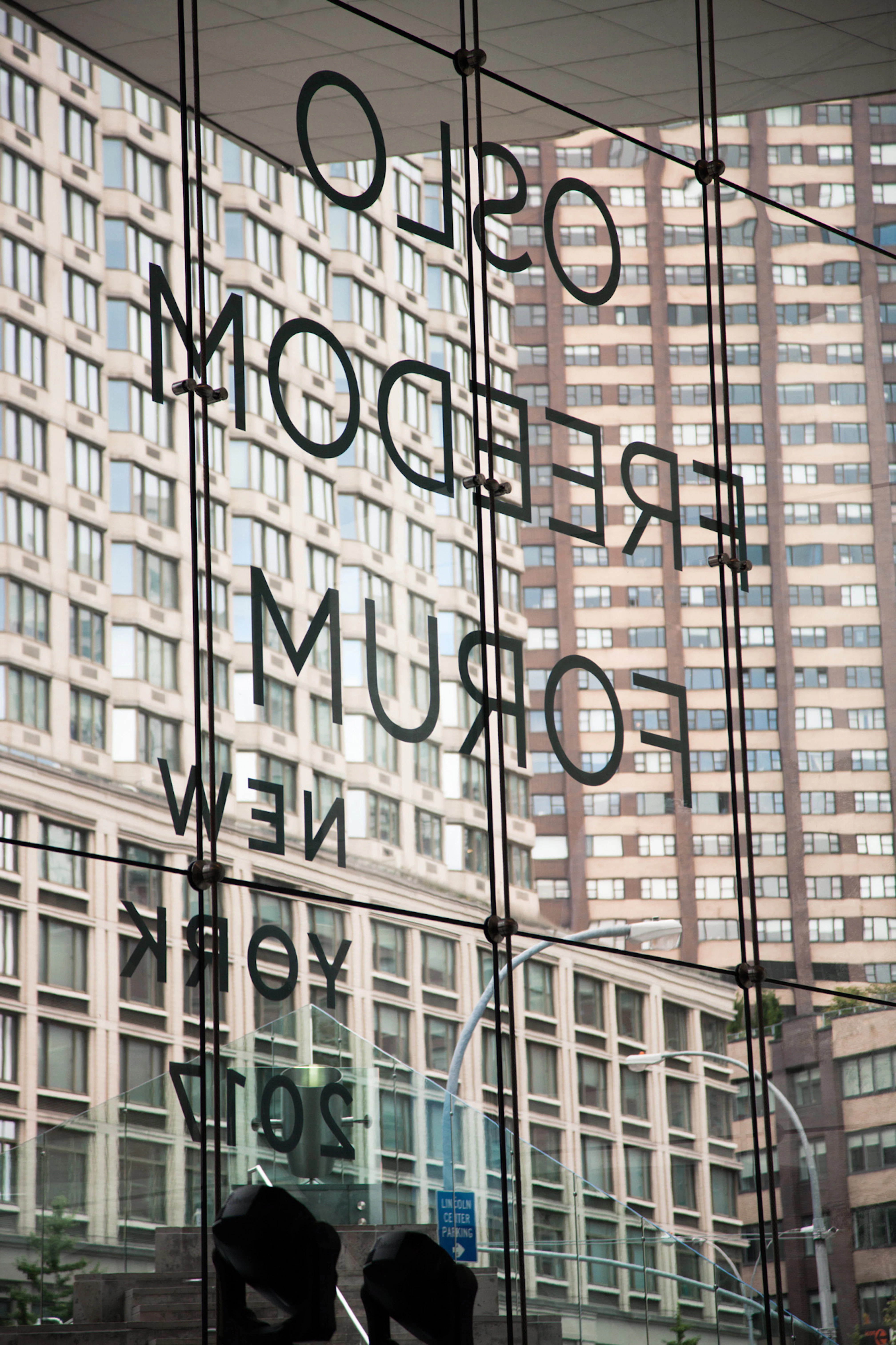 Image shows the words OSLO FREEDOM FORUM NEW YORK 2017 in decal letters on a window. The words are backwards because it is taken from inside the building. Through the window are multi-story city buildings. 