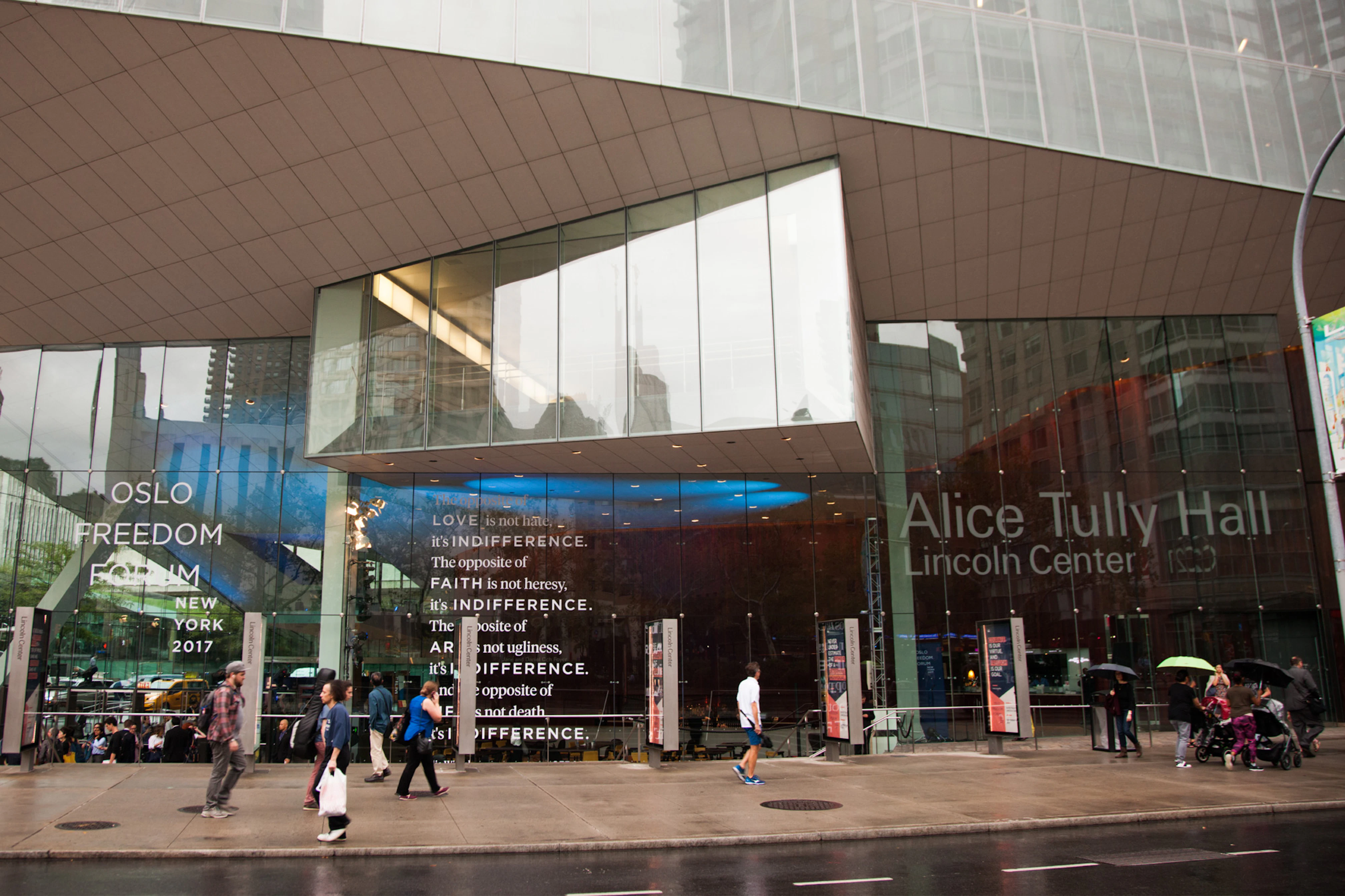 Image shows a contemporary building with glass-paneled walls. Words on the building's windows read OSLO FREEDOM FORUM on the left and Alice Tully Hall Lincoln Center on the right. There are people walking around in front of the building. 