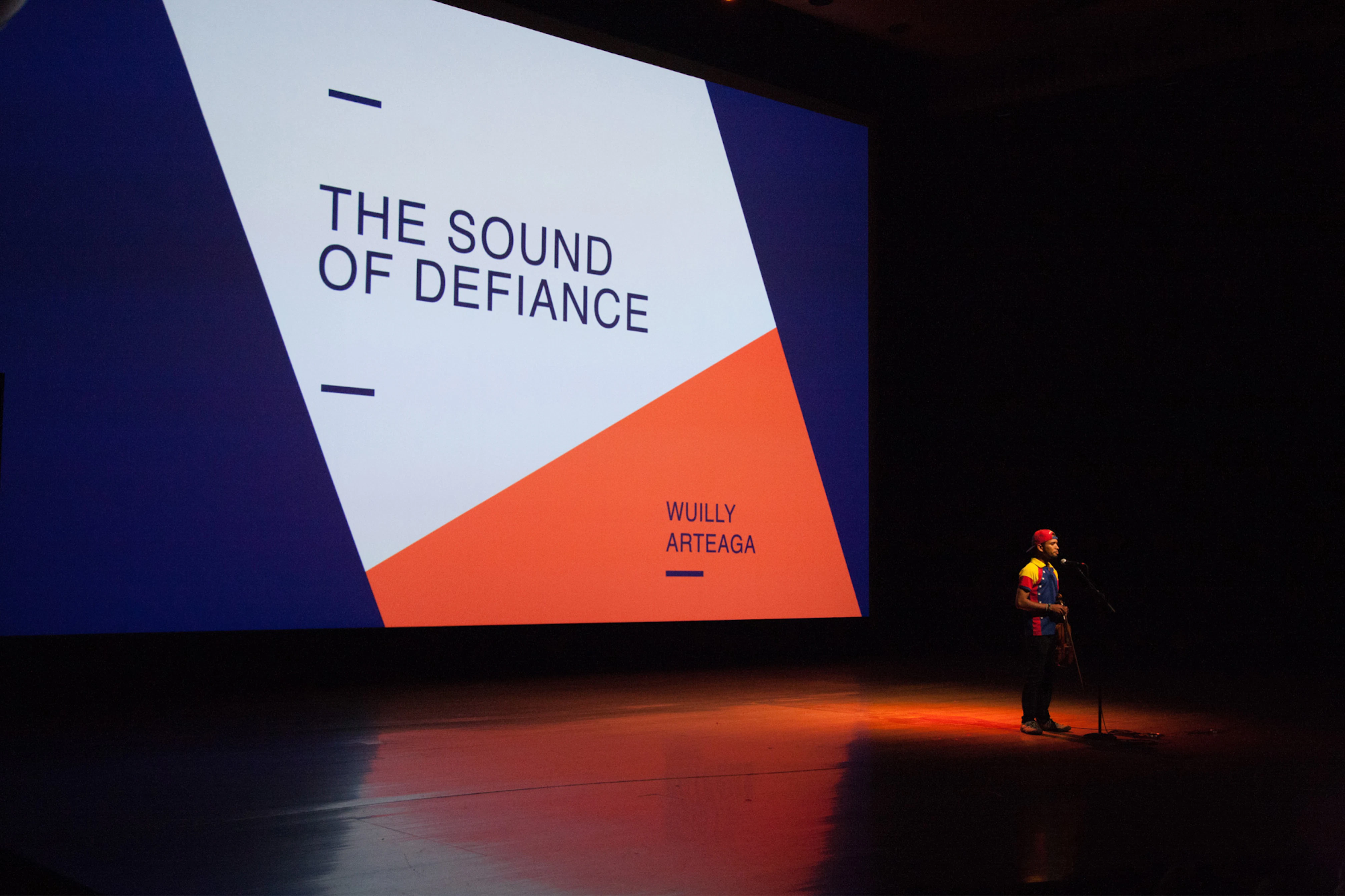 Image shows a person standing on a stage next to a microphone. Behind the person is a large screen with a slide displayed that says THE SOUND OF DEFIANCE in navy blue letters on a white and orange geometric shape. 