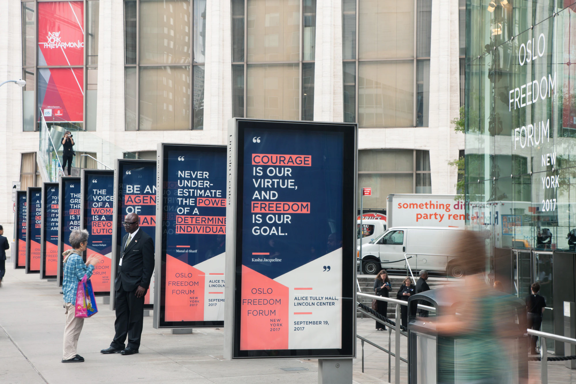 Image shows a row of outdoor signage in a city environment. The signage is navy blue, coral, and white, and there are people stopped nearby talking and others walking past.
