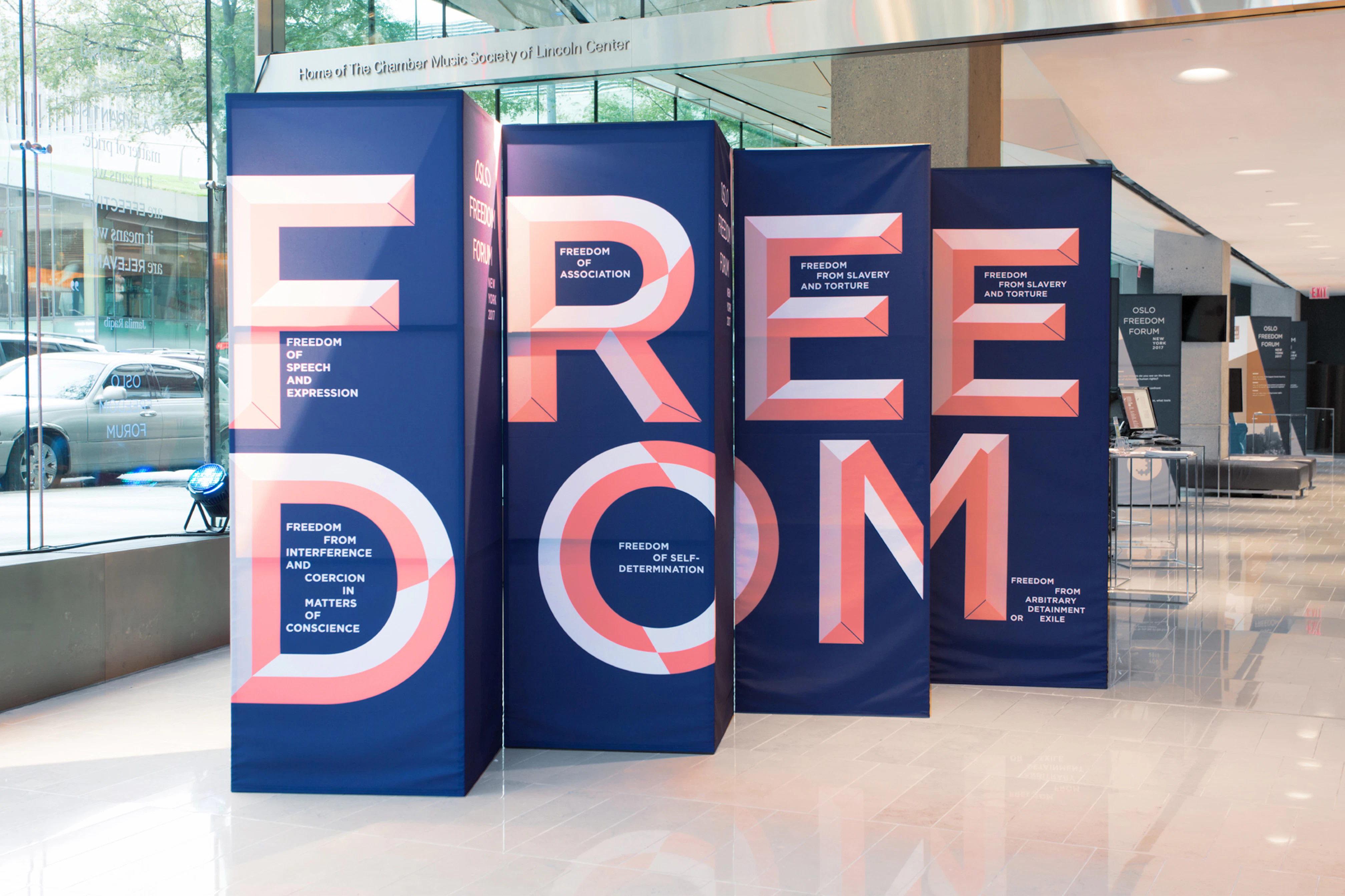 Image shows a set of 4 rectangular, navy blue 3D installations that have the word FREEDOM in large letters on them. They are sitting indoors on a white glossy floor next to a window. 
