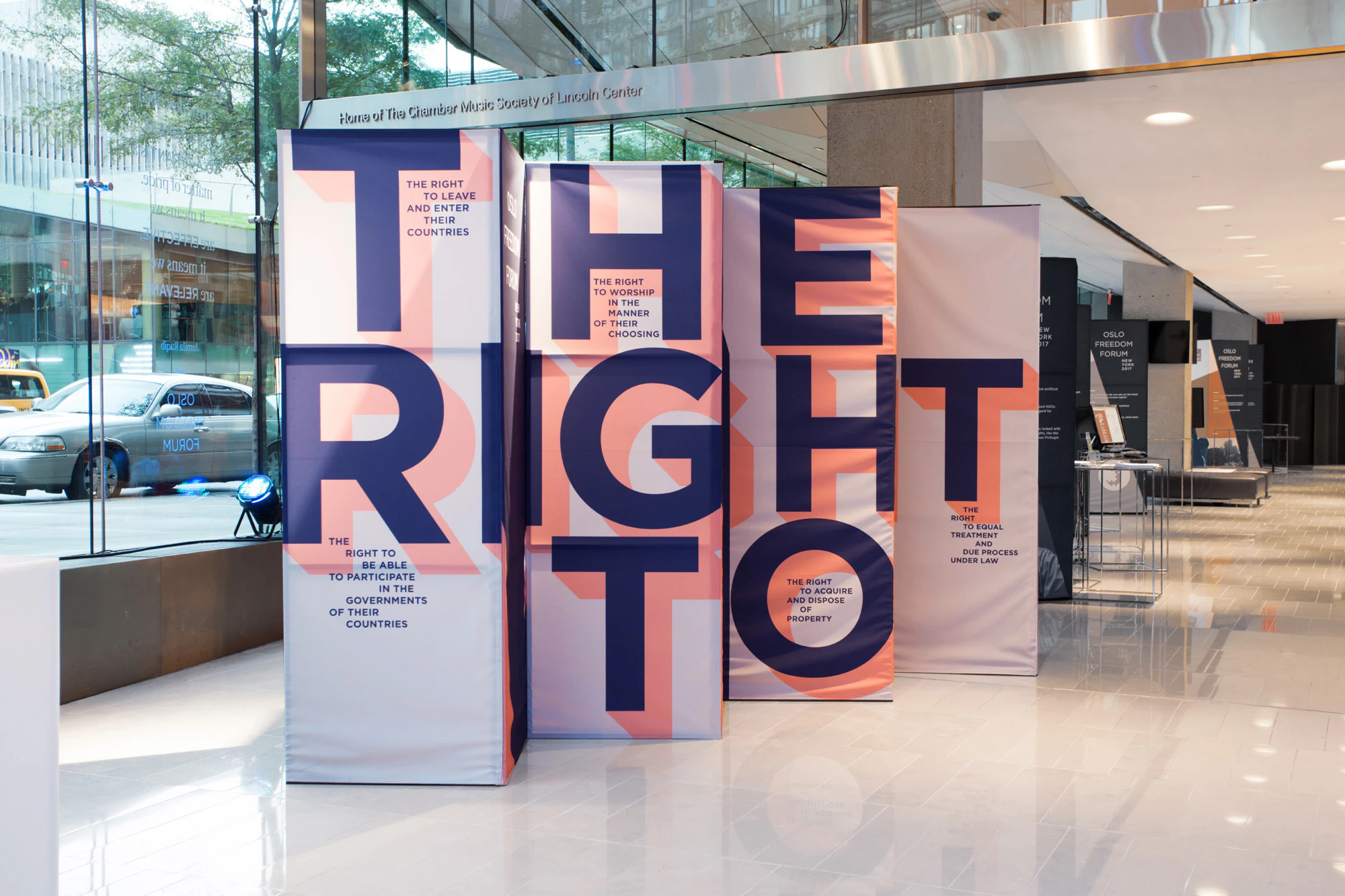 Image shows 4 rectangular 3D installations. Together they show the phrase THE RIGHT TO in navy blue letters with an orange shadow on a light gray background. The installations sit on a glossy white floor inside a building near a window.
