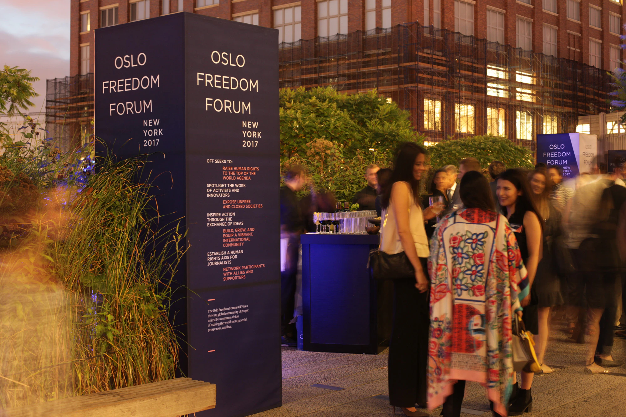 Image shows people gathered in an outdoor space surrounded by buildings and plants. There is a rectangular 3D navy blue installation that reads OSLO FREEDOM FORUM NEW YORK 2017 on it followed by paragraphs of smaller text in white and orange.