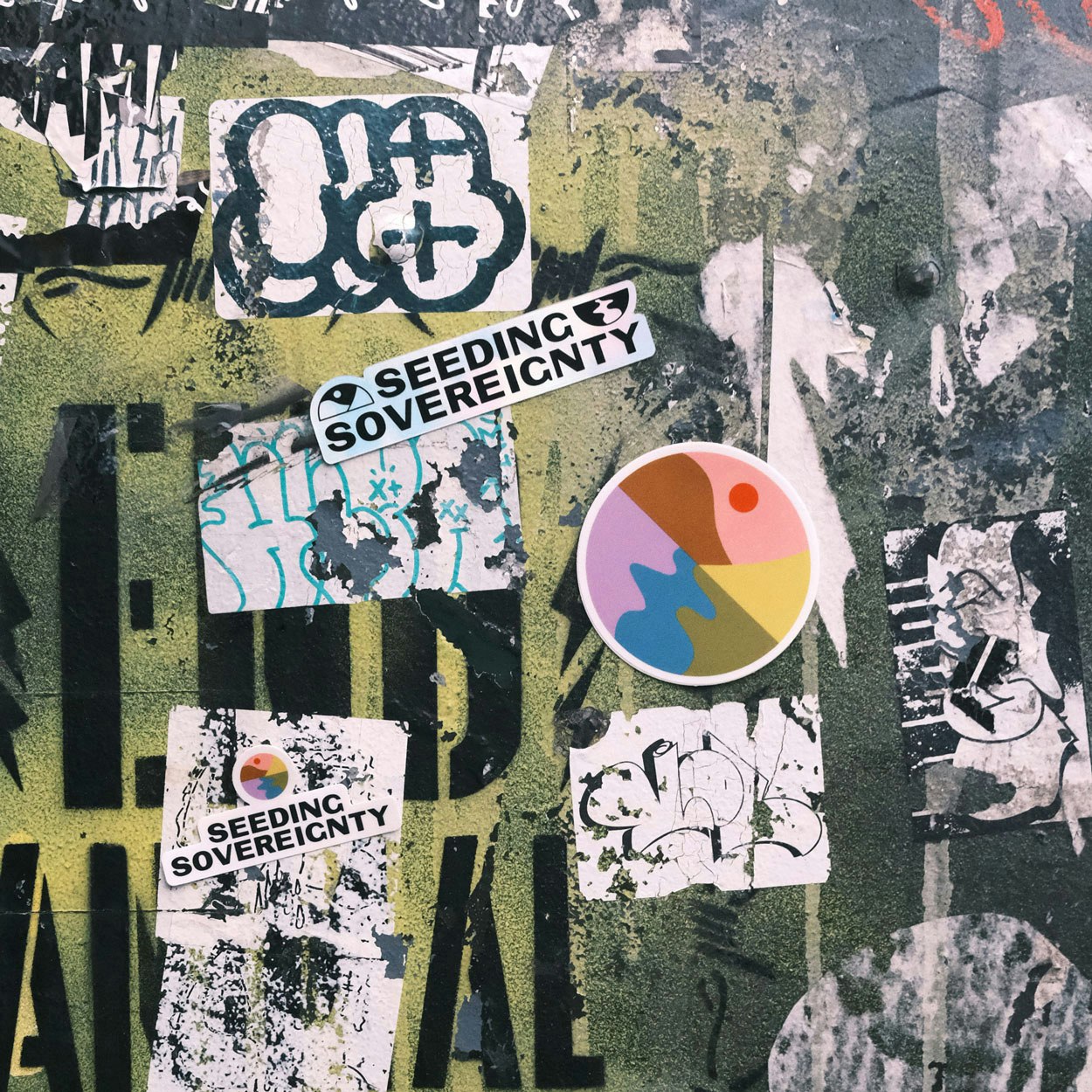 Image shows Seeding Sovereignty stickers on a surface covered in old peeling stickers and graffiti.