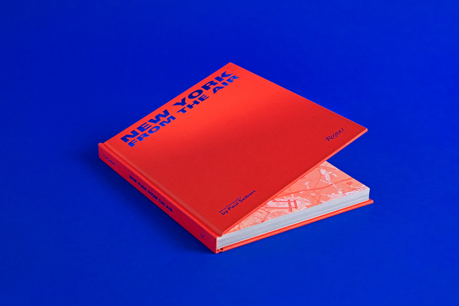 Photo showing the red book almost closed but open enough to get a glims of the pattern inside of it. The title NEW YORK FROM THE AIR is printed in cobalt blue foil and the book is resting on the same color seamless.