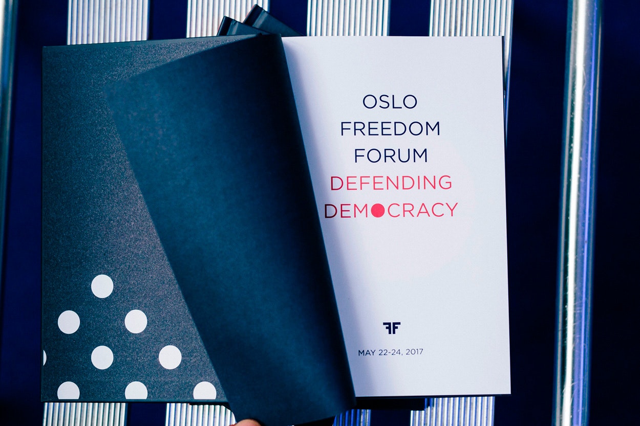 Image shows a book open on a white and blue striped surface with a navy blue page flipping to reveal a white page that says, "OSLO FREEDOM FORUM DEFENDING DEMOCRACY"