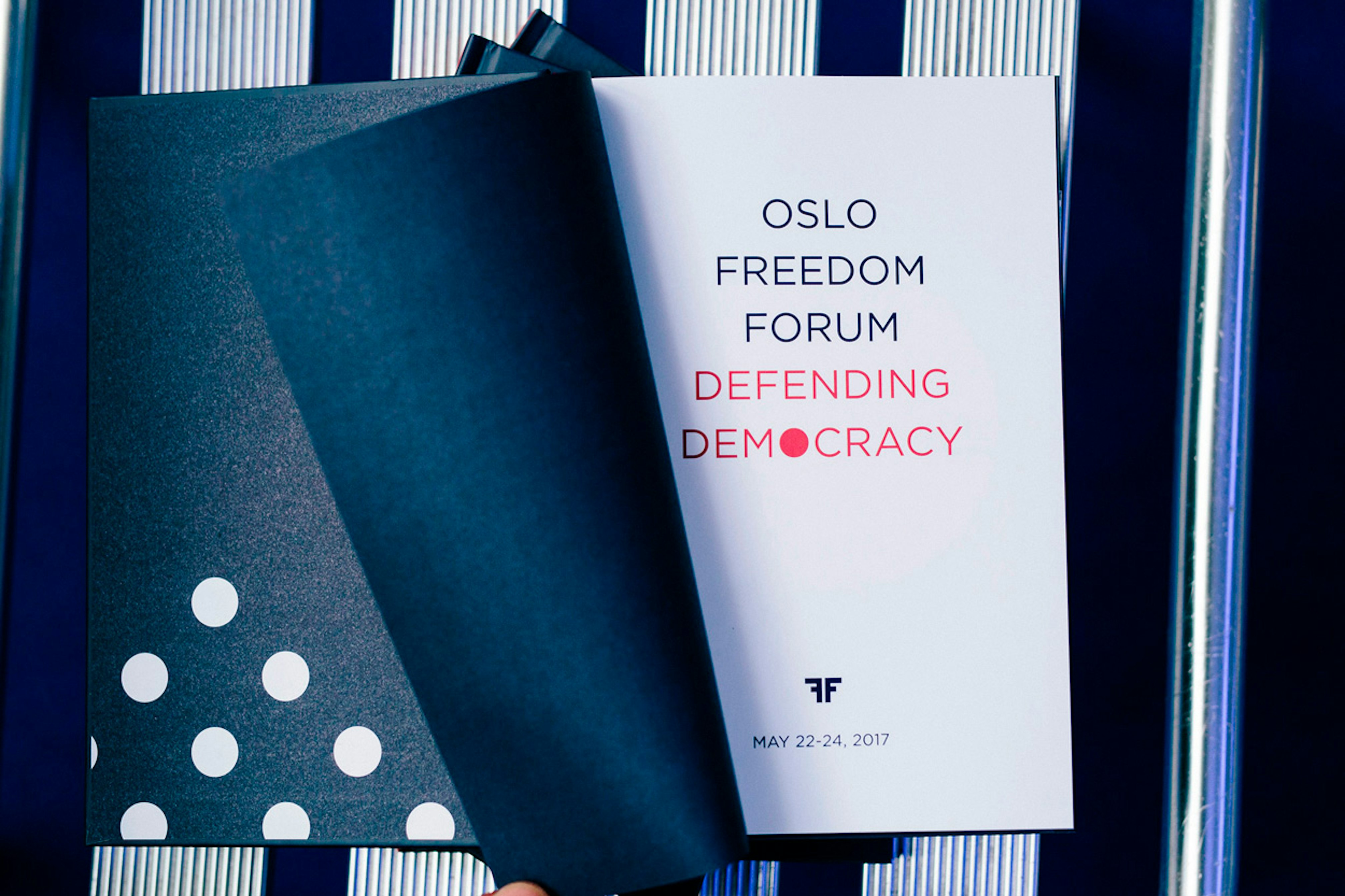 Image shows a book open on a white and blue striped surface with a navy blue page flipping to reveal a white page that says, "OSLO FREEDOM FORUM DEFENDING DEMOCRACY"