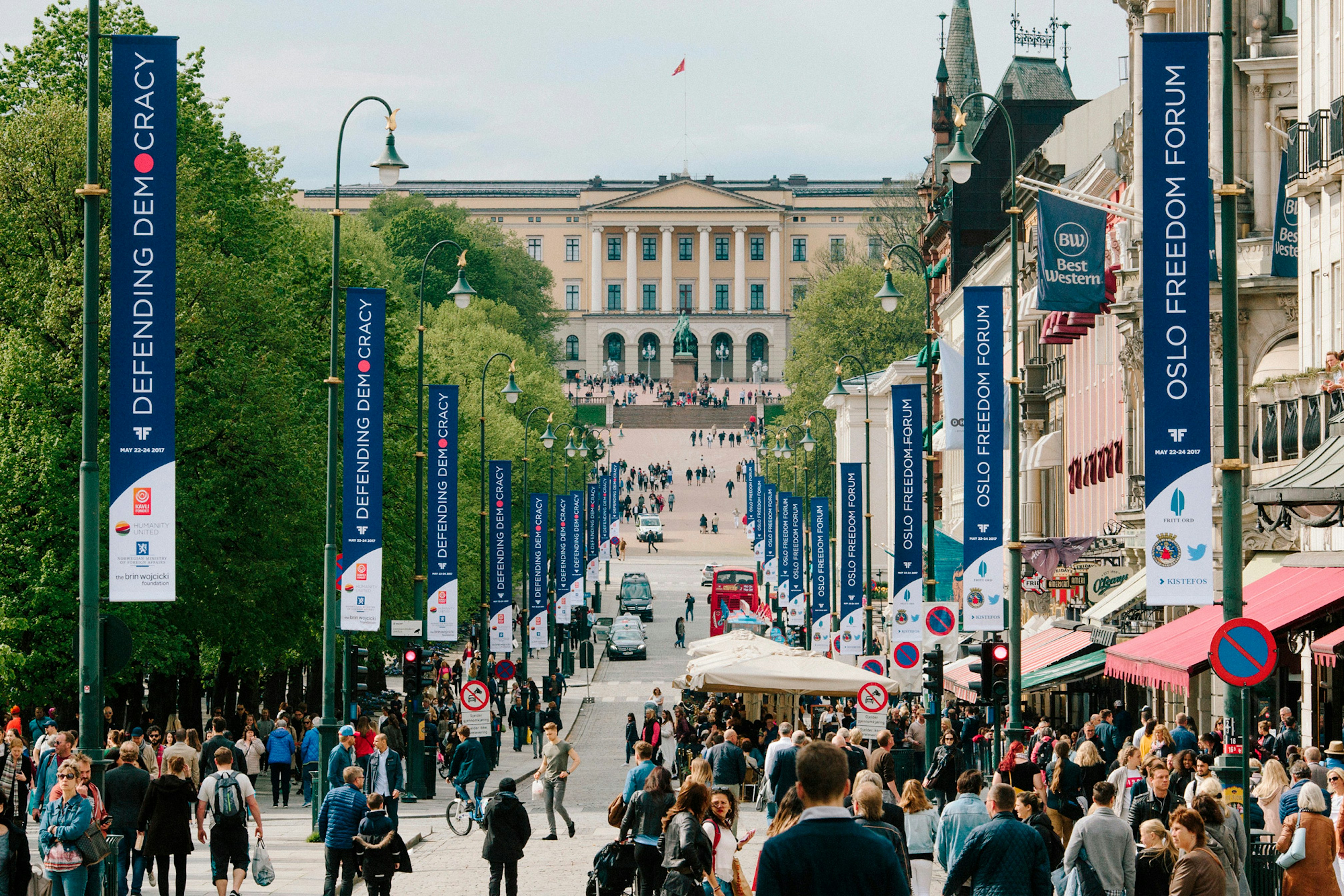 Image shows a road leading to a large building that is lined in navy blue Oslo Freedom Forum banners. The street is filled with people walking in the direction away from the camera and lined in trees. 