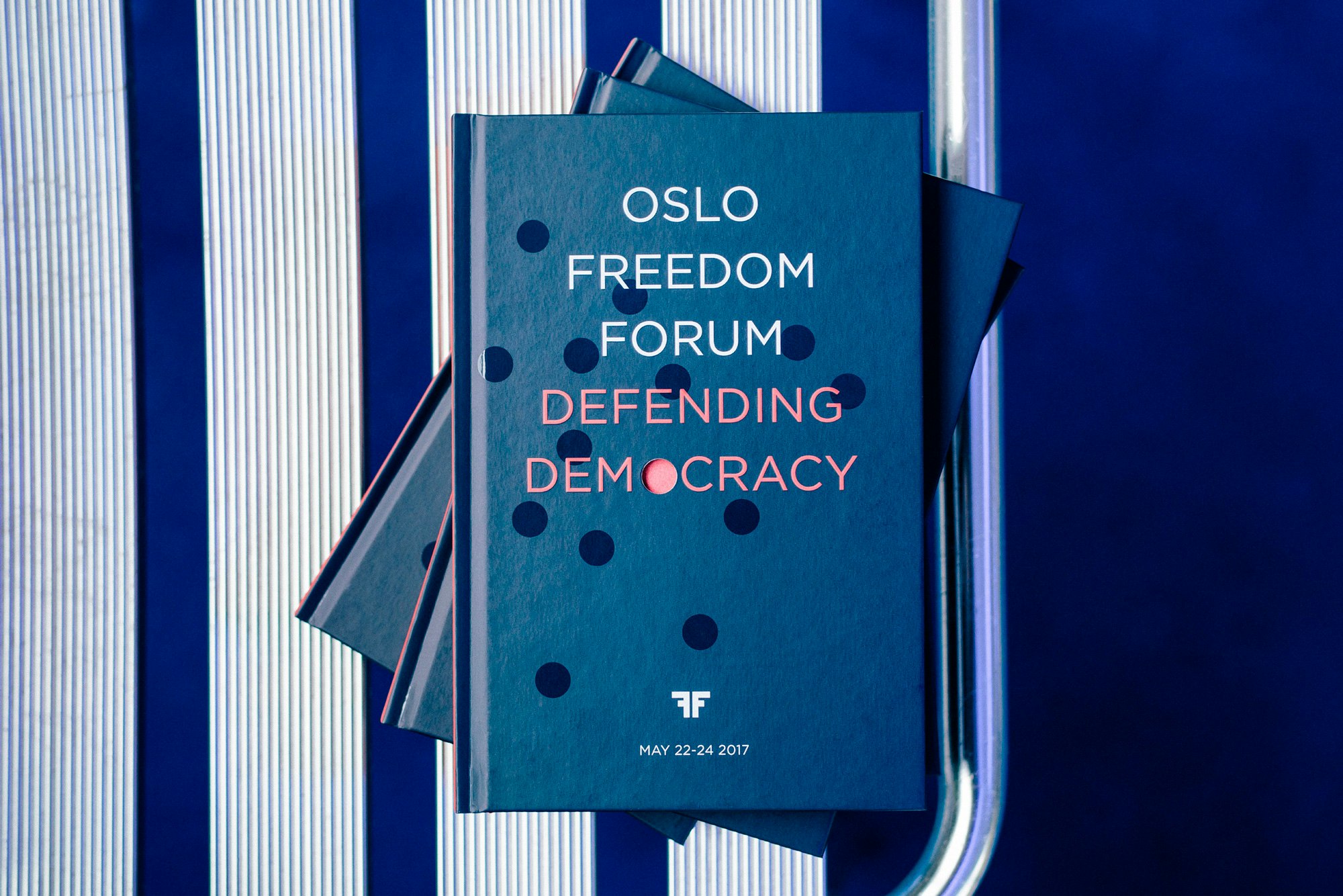 Image shows a book open on a white and blue striped surface with a navy blue page flipping to reveal a white page that says, "OSLO FREEDOM FORUM DEFENDING DEMOCRACY"