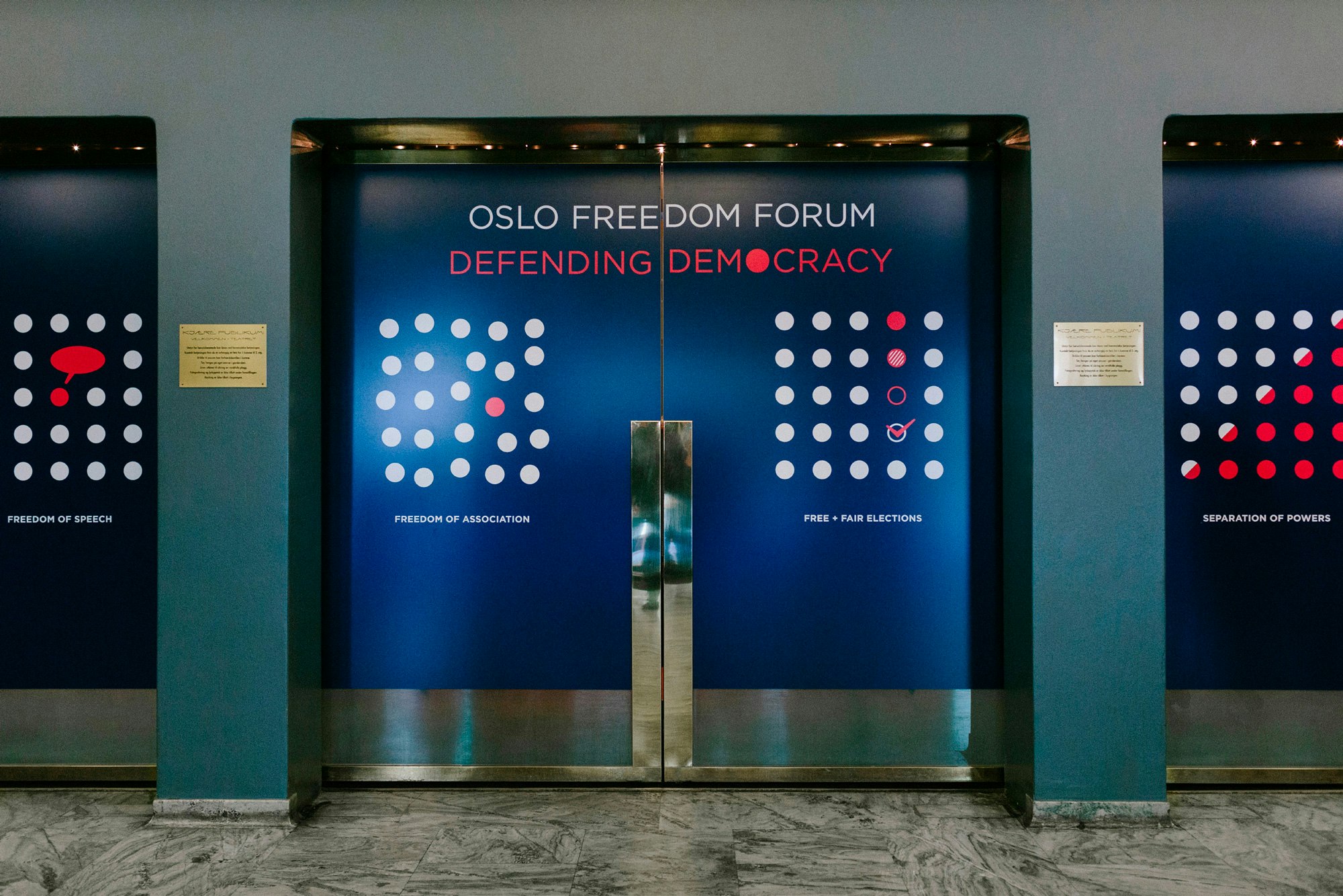 Image shows Oslo Freedom Forum branding on elevator doors. They are blue with OSLO FREEDOM FORUM DEFENDING DEMOCRACY written on them, along with white and coral dots.