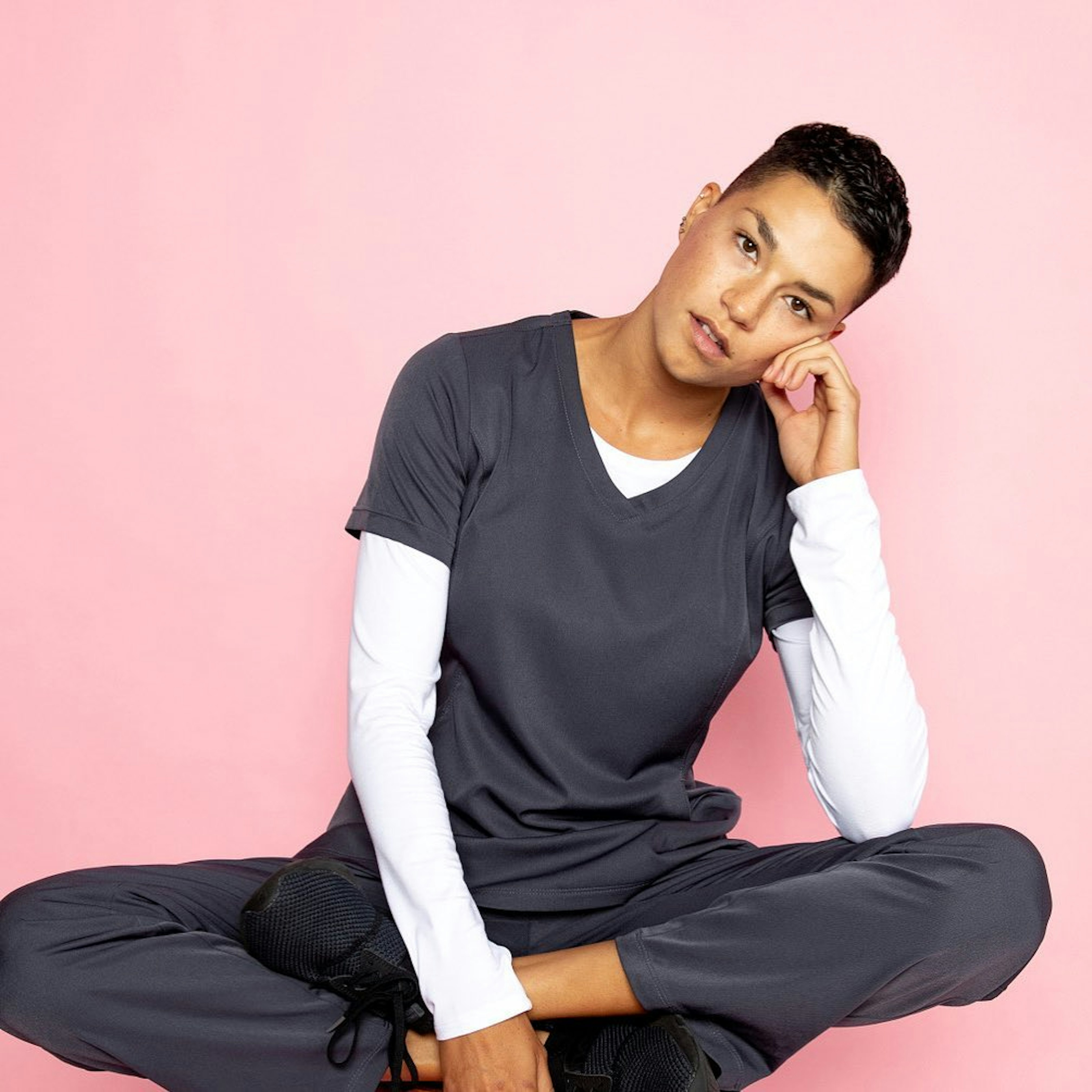 A person sitting down on a floor wearing dark gray scrubs with white long sleeves inside. The background color is light pink.