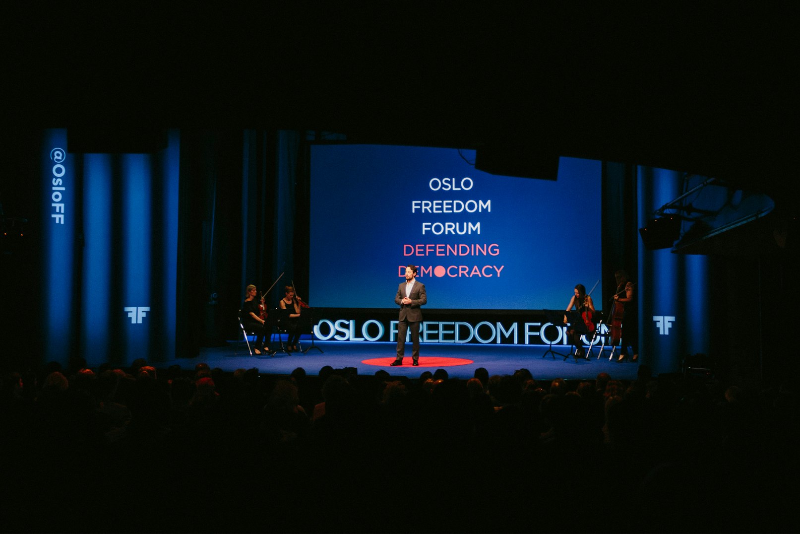Image shows a person on a blue stage with a blue screen behind them that reads, "OSLO FREEDOM FORUM DEFENDING DEMOCRACY. Two people sit in chairs on either side of the person standing in the middle of the stage. 
