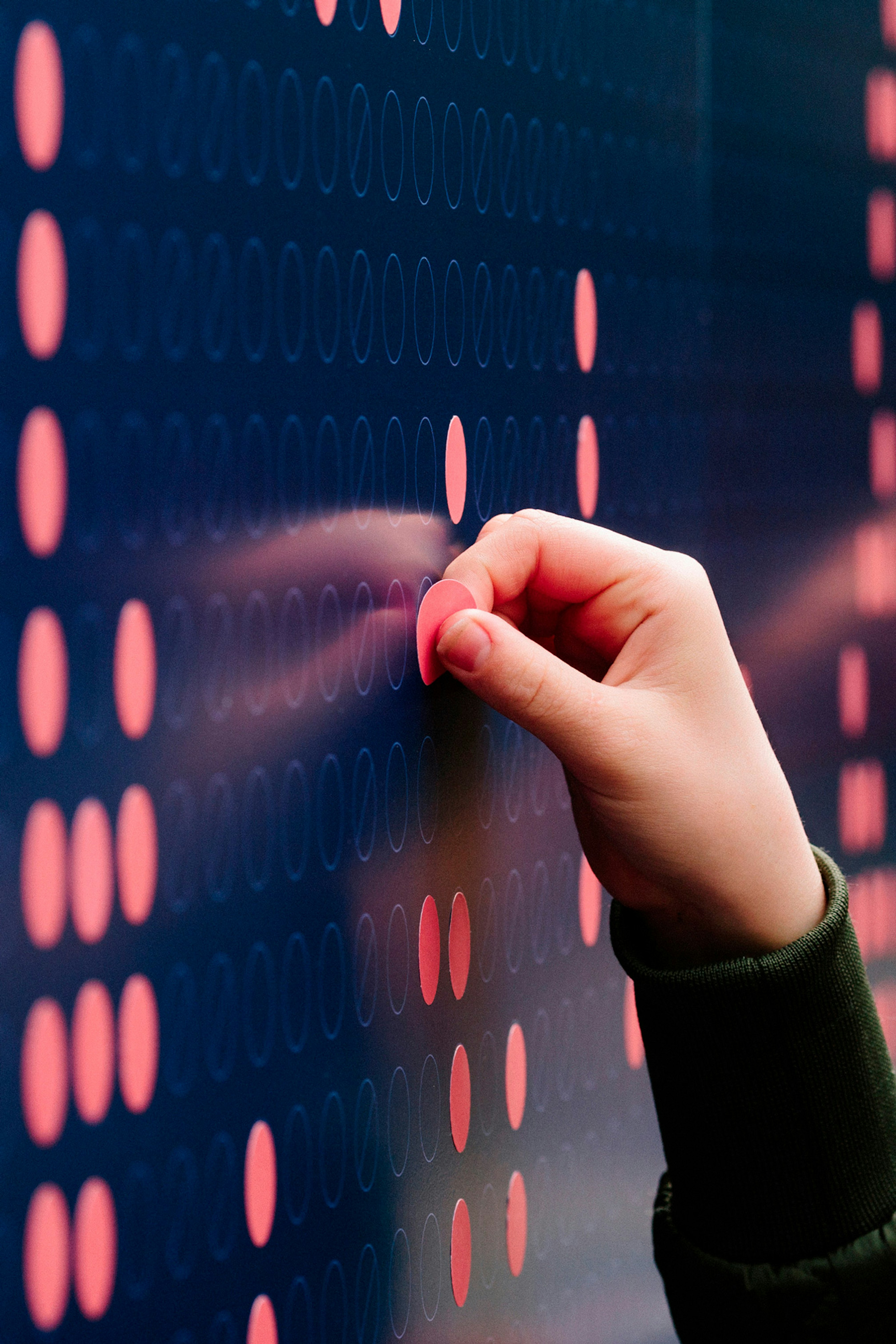 Image shows a close-up of a hand with pale skin placing a coral-red dot sticker on a navy blue wall. 