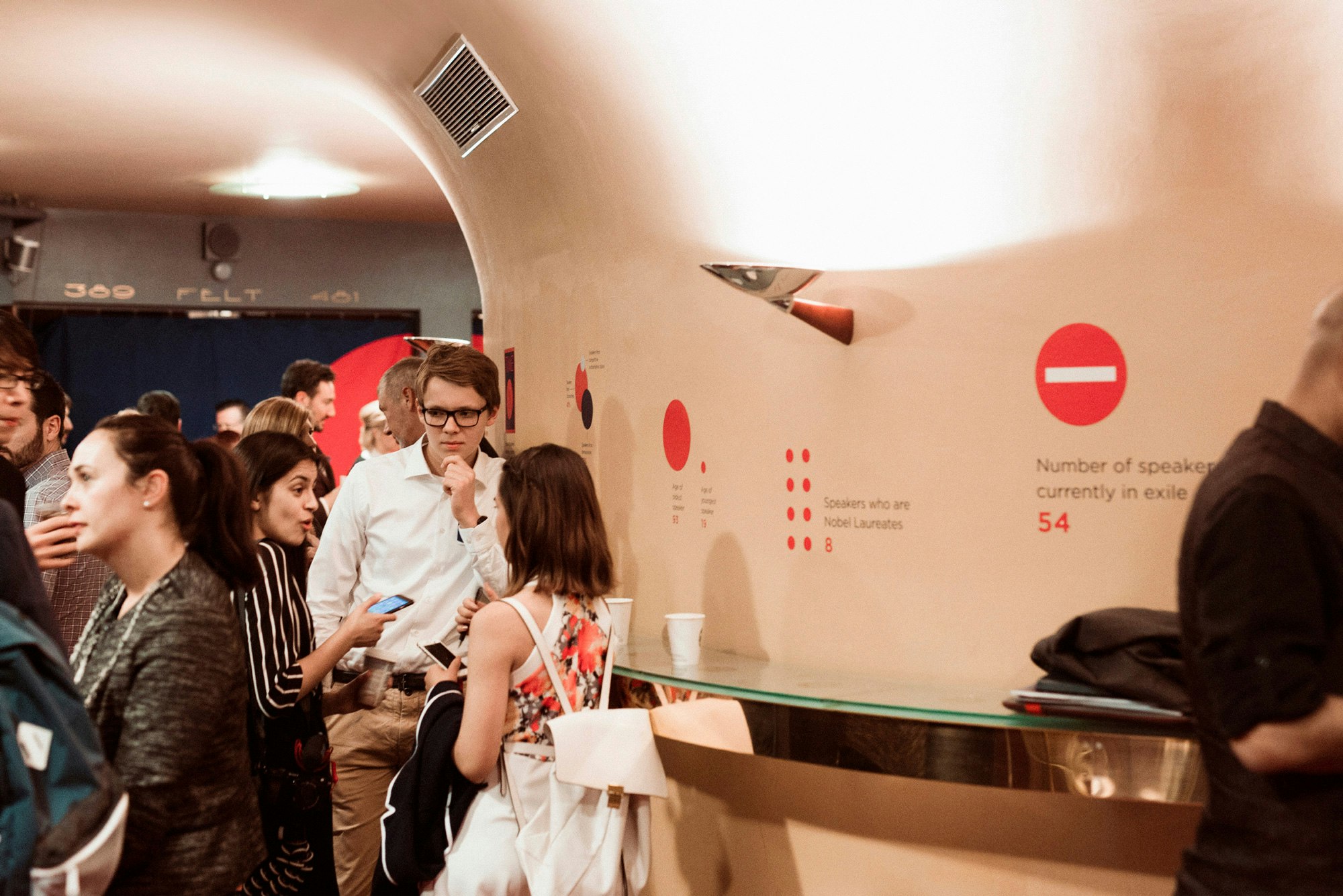 Image shows people gathered and talking in a space with white walls and curved ceiling. There are red infographics on the walls of the space.