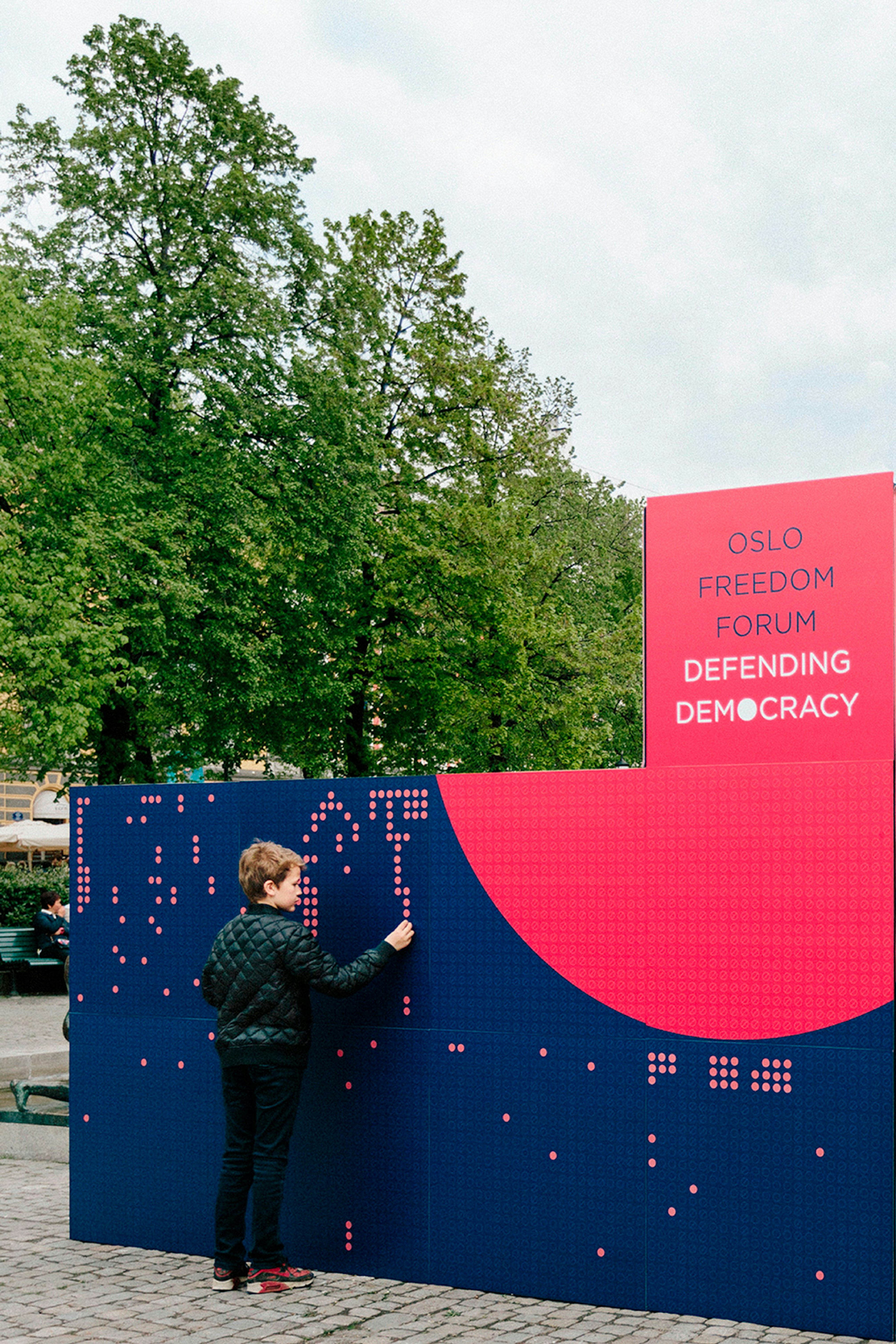 Image showing a kid in the park in Oslo putting a round red sticker on an activation wall that when all the stickers are placed will say DEFEAT DICTATORSHIP