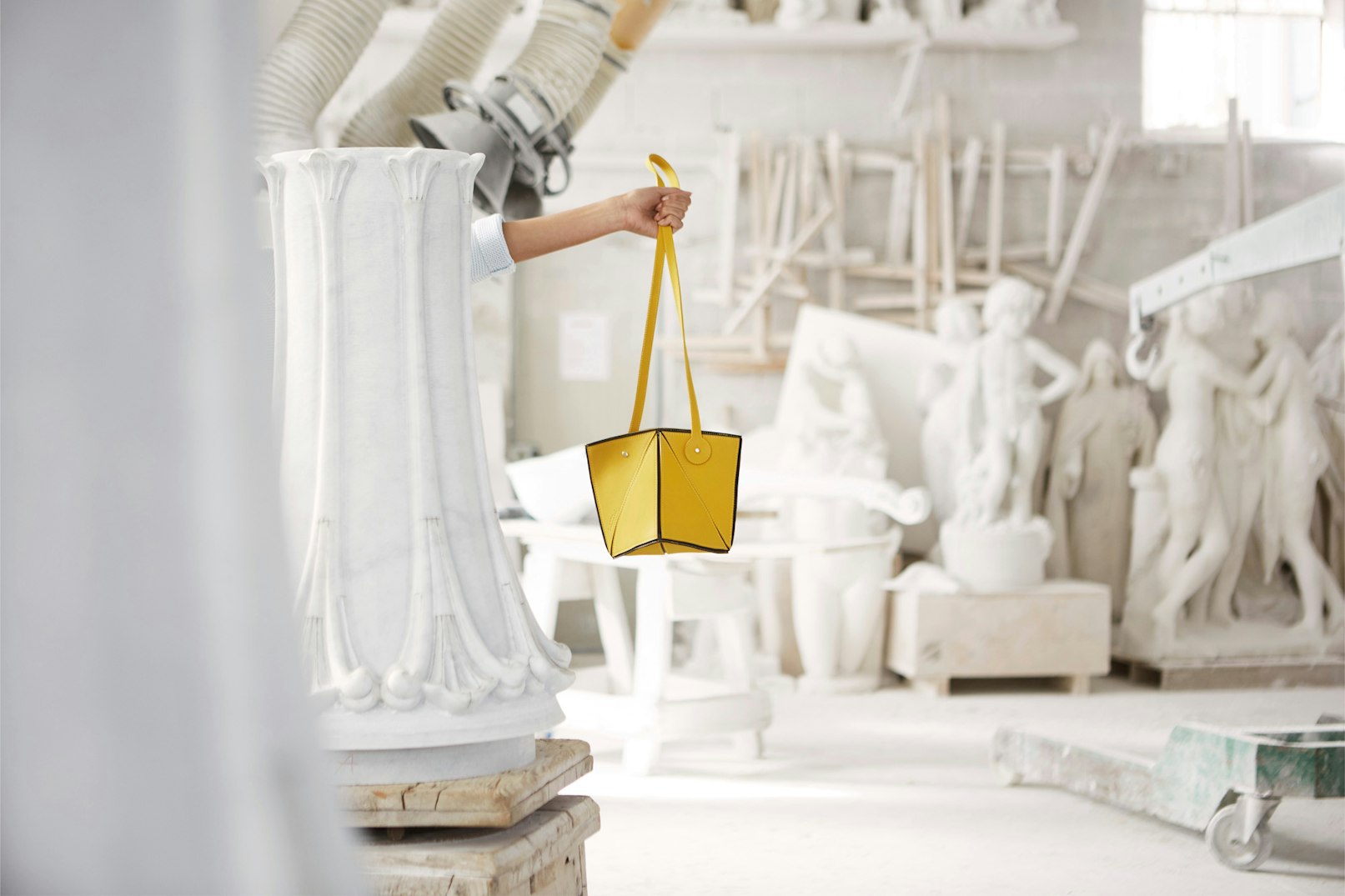 Image shows an arm reaching out from a piece of a white column, in a room full of white sculpture fragments, holding a geometric, yellow leather handbag. 