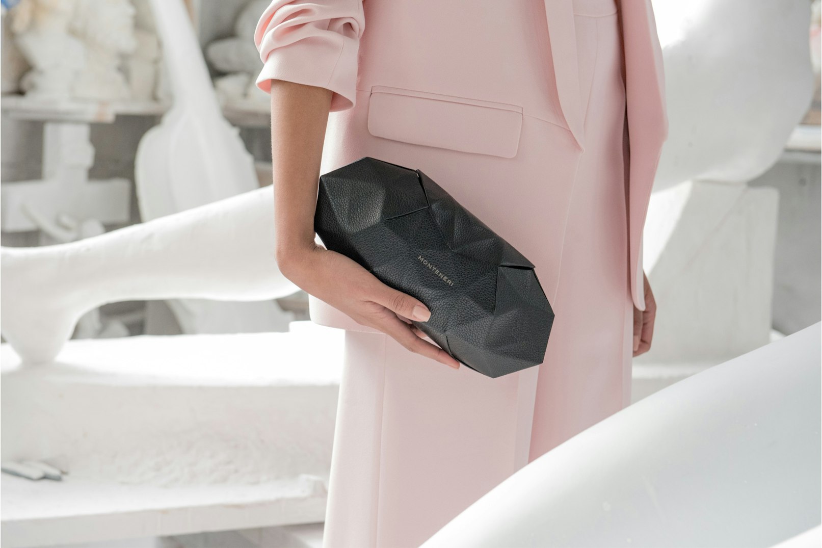 Image shows the middle portion of a person in a light pink suit surrounded by white sculpture fragments, holding a geometric black leather clutch bag. 
