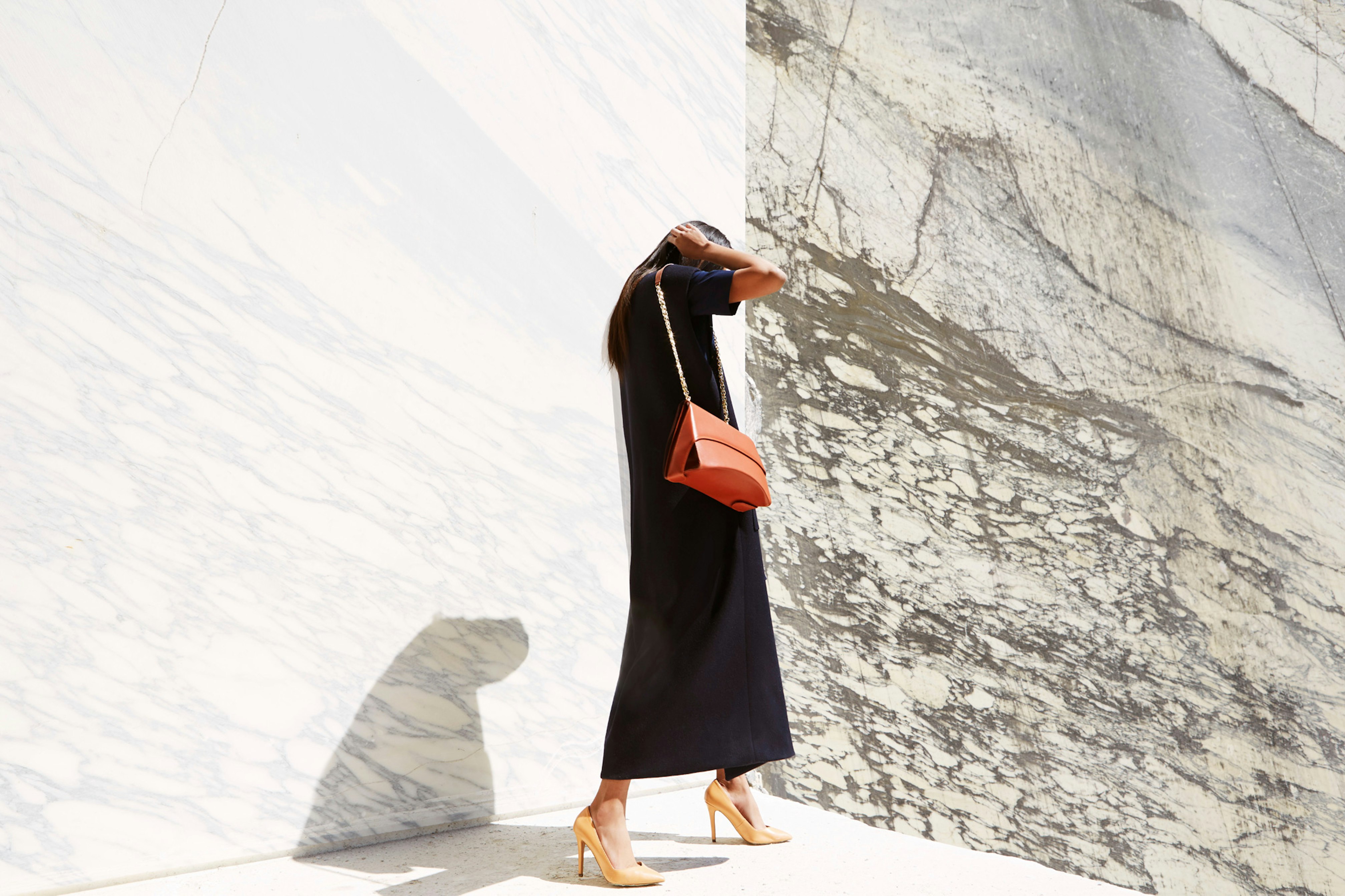 Image shows a person in a long black dress against a marble background wearing an orange-toned leather bag and beige heels. They are touching their dark hair, and you do not see their face. 