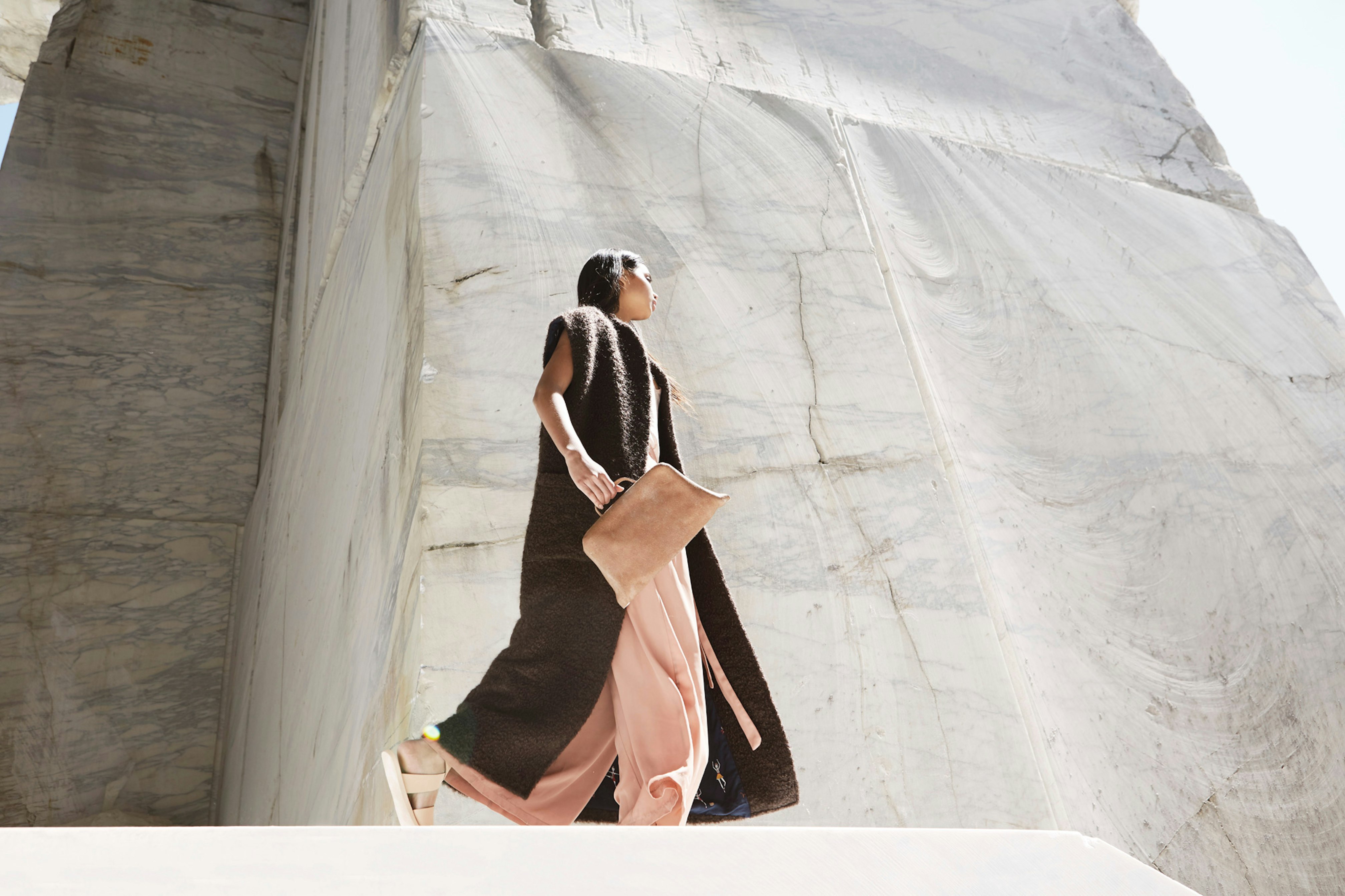 Image shows a person walking in a blush pink jumpsuit, a long duster with short sleeves, and off-white sandals while holding a light beige leather bag. 