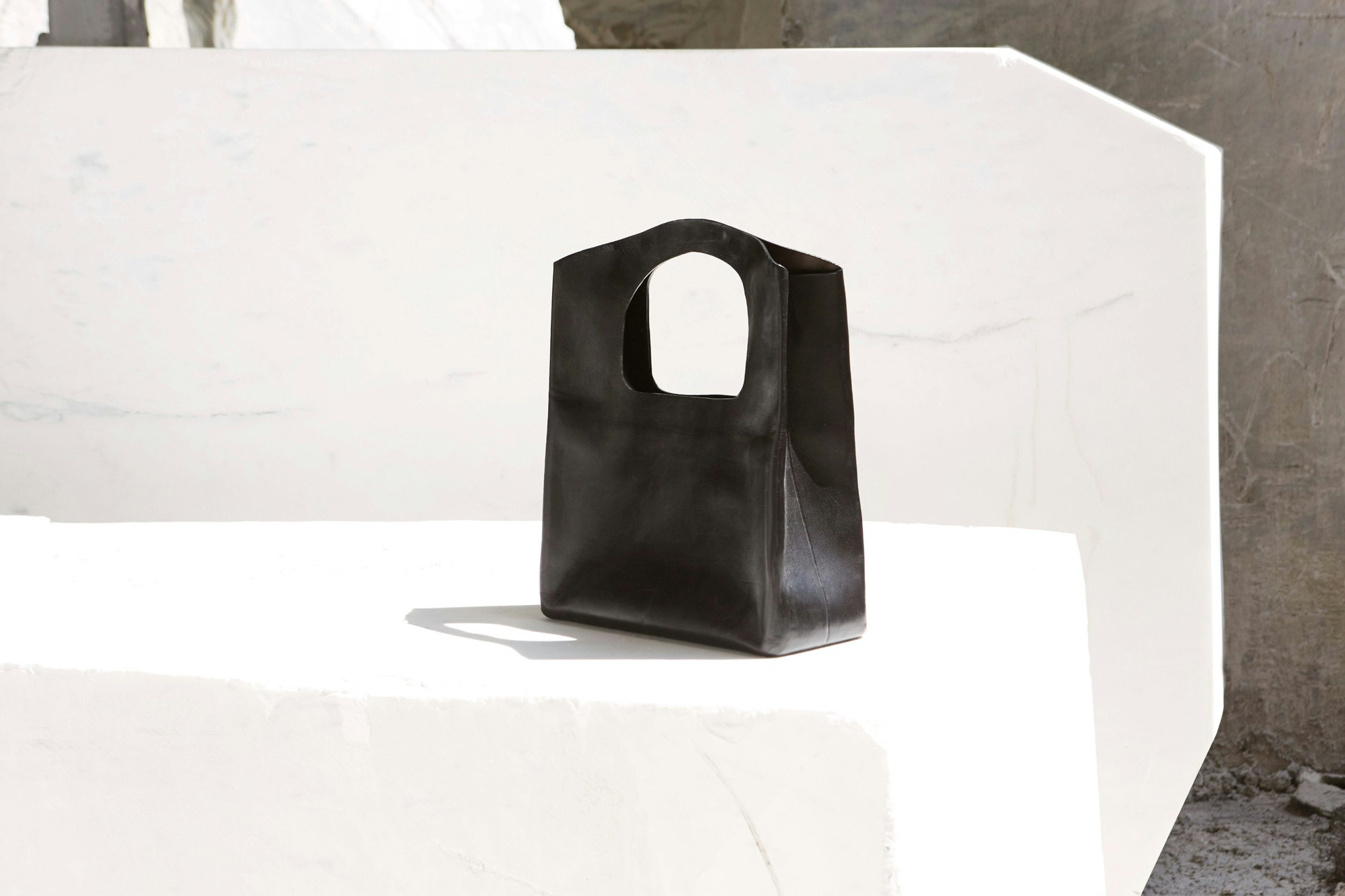 Image shows a geometric black handbag sitting on a bench-shape of white marble.