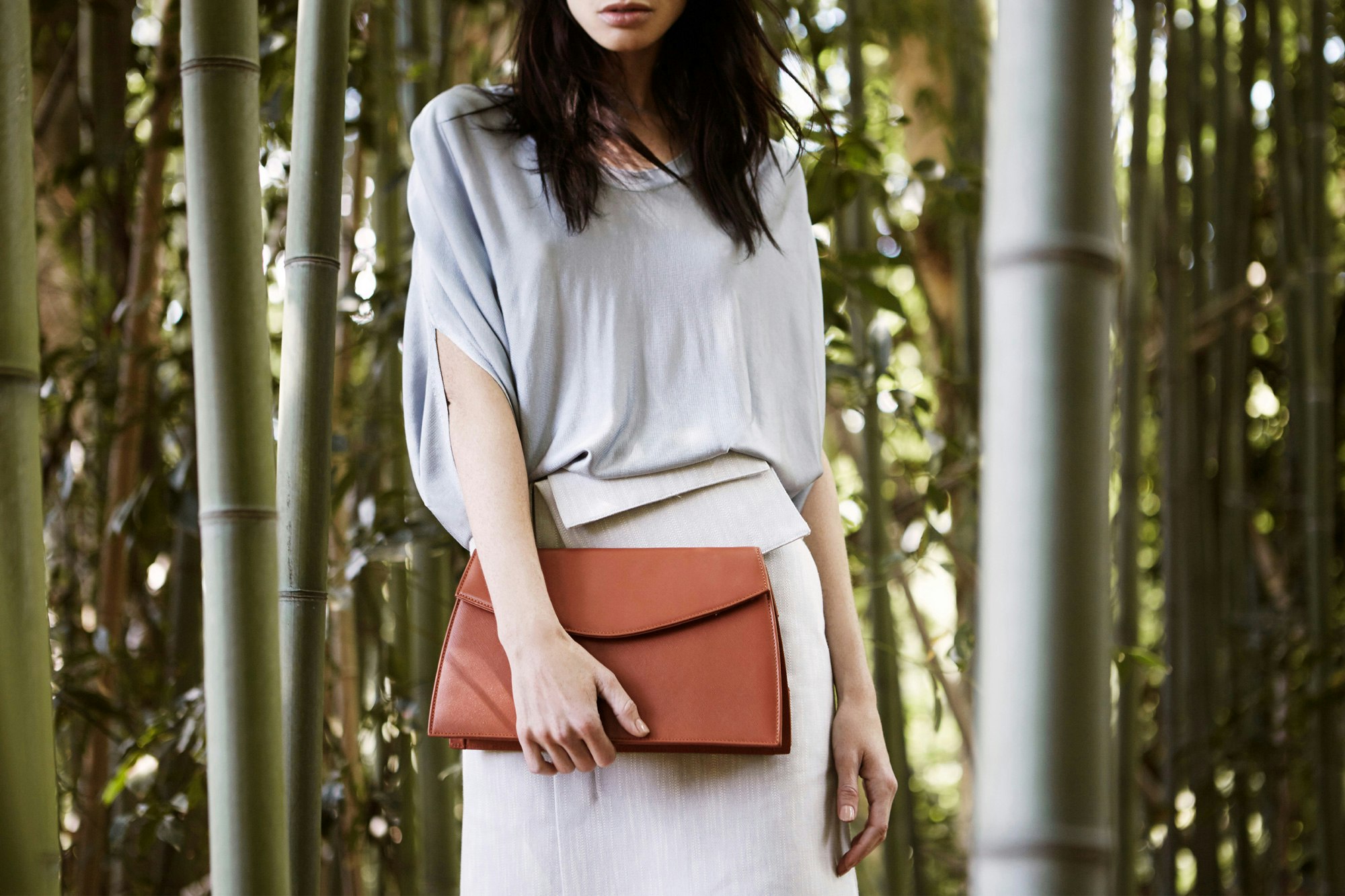 Image showing the middle portion of a person in a light grey outfit holding a ochre leather clutch standing in a forest of bamboo.