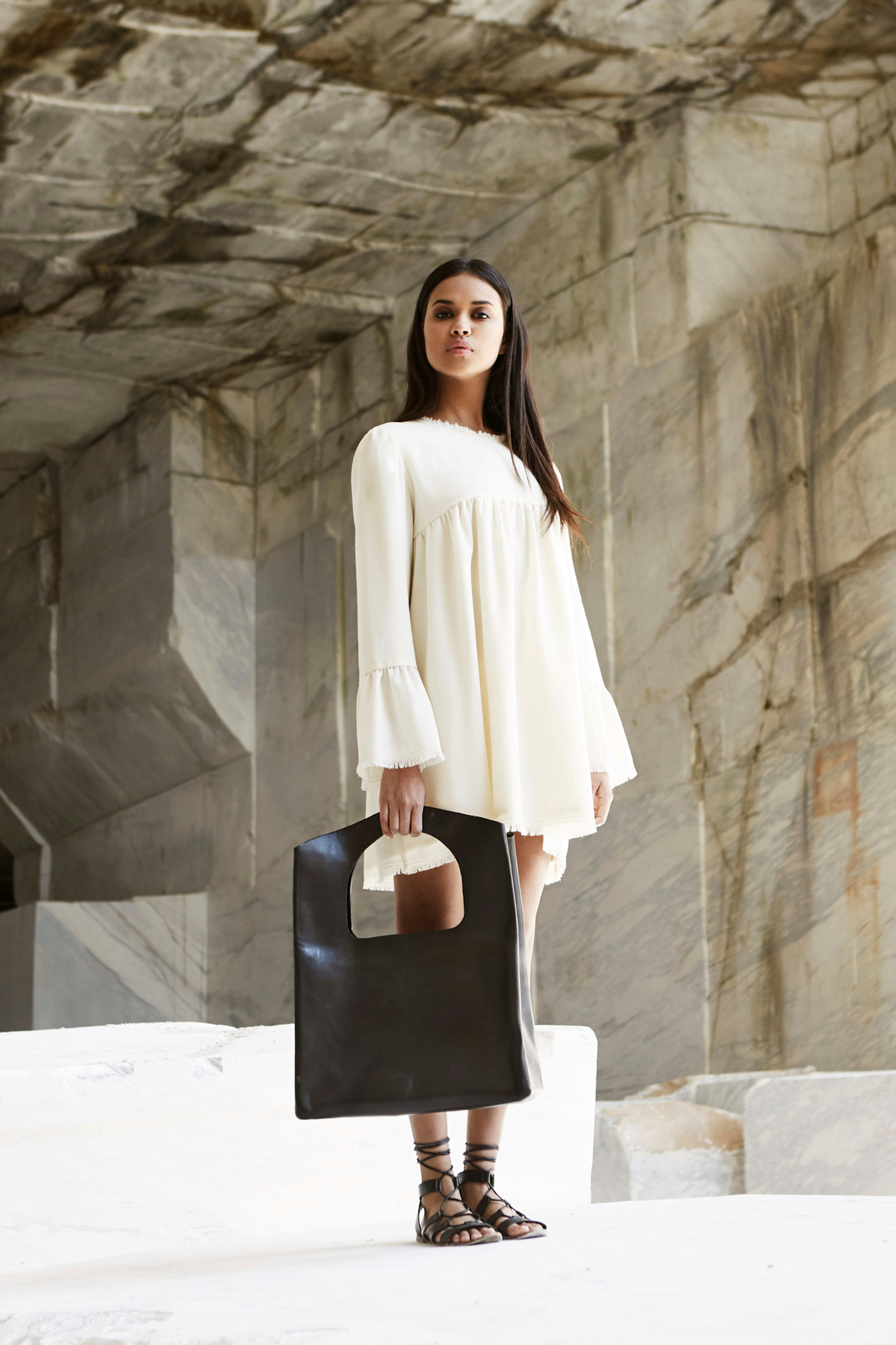 A person in a short off-white dress holds a large black leather handbag and stands surrounded by marble slabs. 