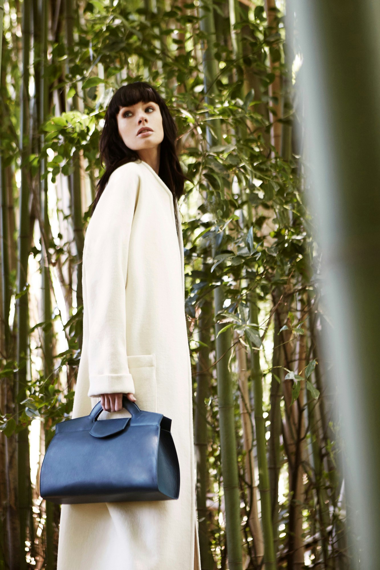 Image shows a person with dark hair wearing a long off-white jacket holding a navy leather bag and standing in a forest of bamboo. They look back with their face toward the camera but their eyes direction beyond the camera.